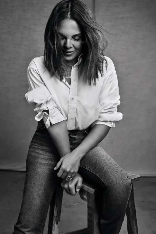 A black and white photo of a woman with shoulder-length hair, wearing a white shirt with rolled-up sleeves and jeans, sitting on a wooden stool with a textured wall background.