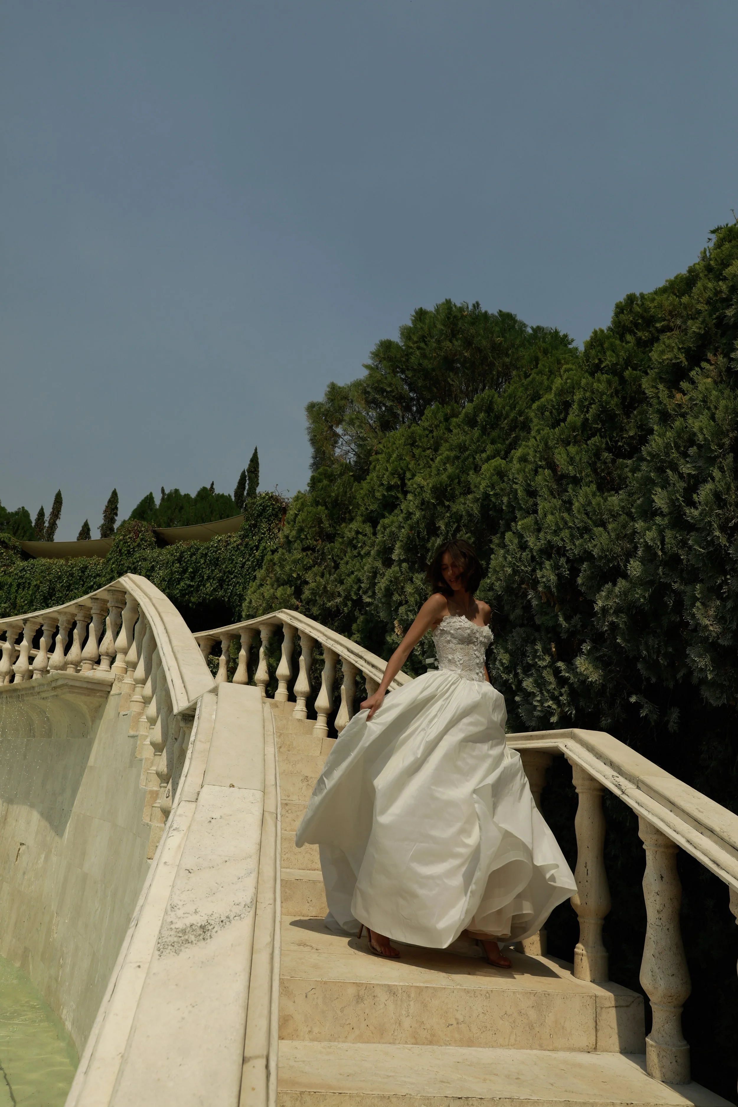 	
Guardiola at New York Bridal Market A woman in a white wedding dress standing on a marble staircase outdoors, surrounded by green trees and clear blue sky.