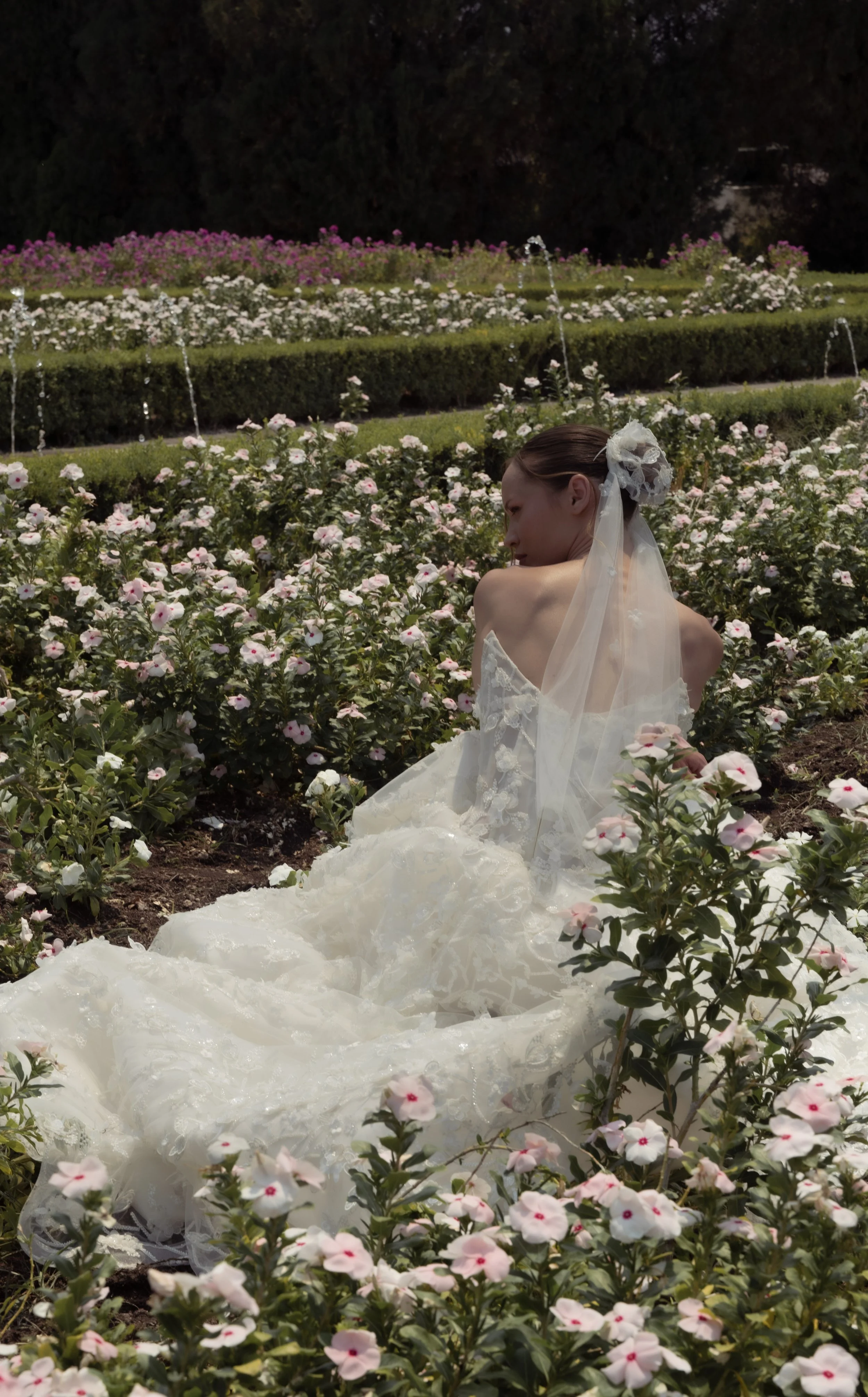 	
Guardiola at New York Bridal Market A bride sitting in a garden filled with white and pink flowers with water fountains in the background, wearing a white wedding dress and veil.