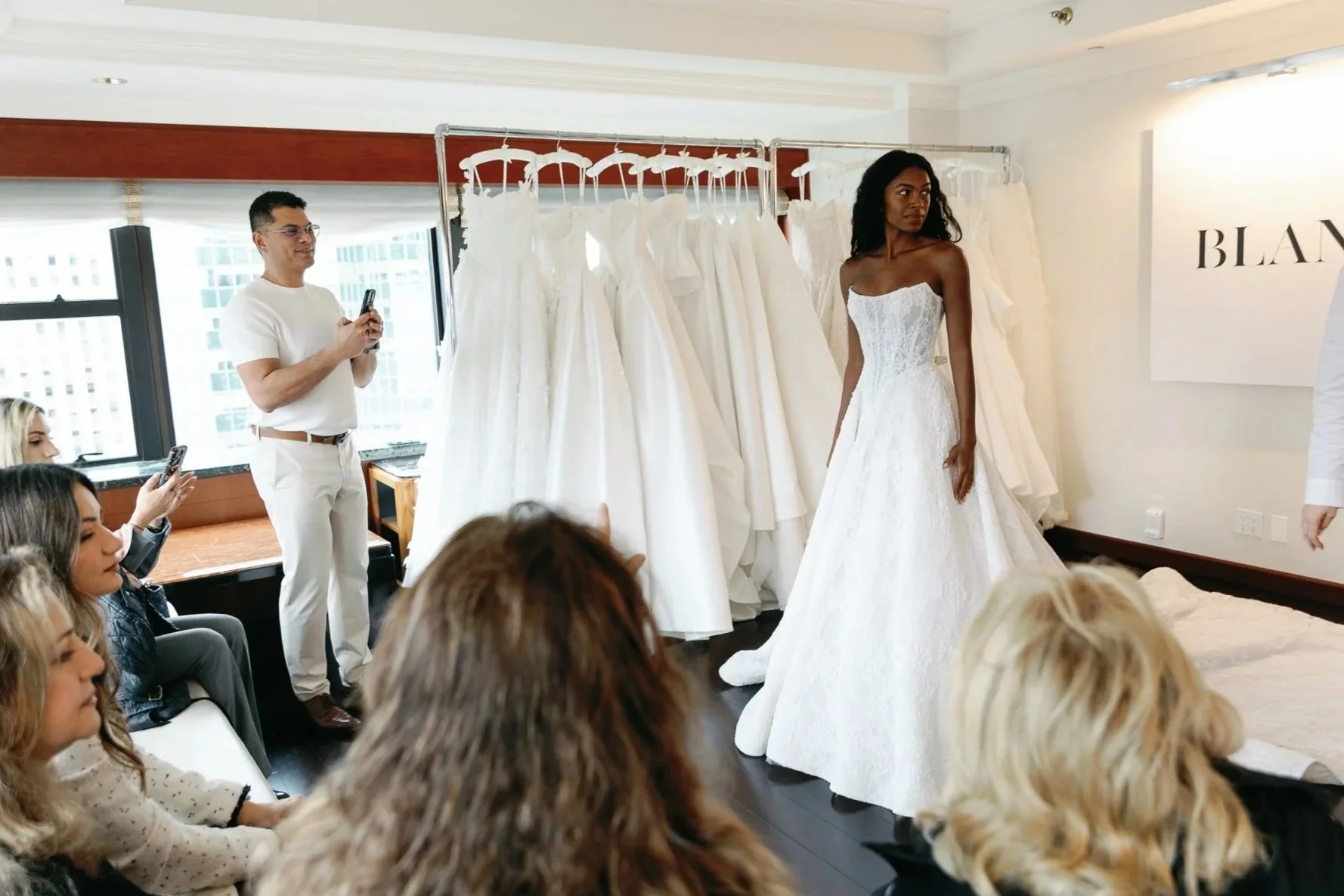 A woman tries on a wedding dress in a boutique, surrounded by friends and staff, with wedding gowns on display behind her.