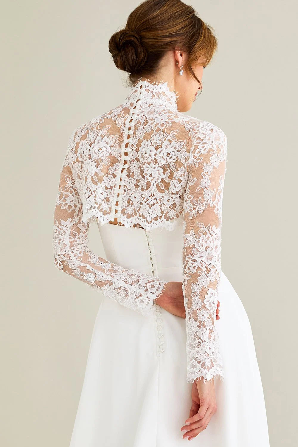 Lea-Ann Belter at New York Bridal Market A woman wearing a white wedding dress with a lace jacket, styled with a bun hairstyle and earrings, standing against a plain background.