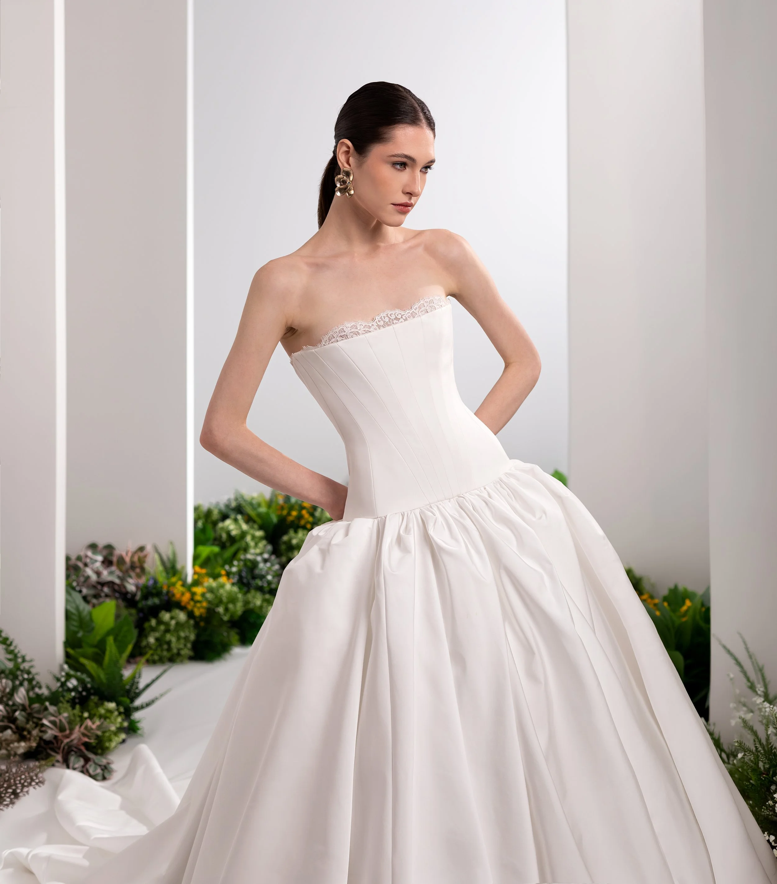Valentini Spose at New York Bridal Market.    A woman in a white strapless wedding gown with lace detail near the top, standing in front of a backdrop with greenery and flowers. She has dark hair pulled back and large gold earrings.