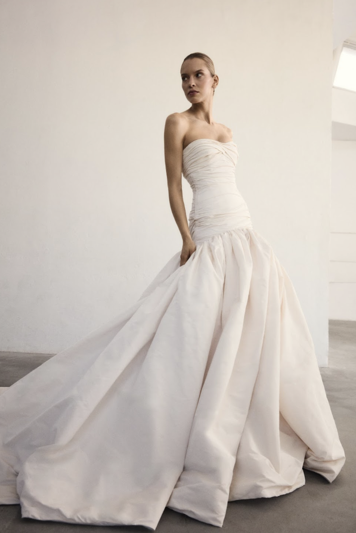 Marmar Halim at New York Bridal Market A woman wearing a strapless, off-white wedding gown with a full, flowing skirt standing against a plain, light-colored wall.