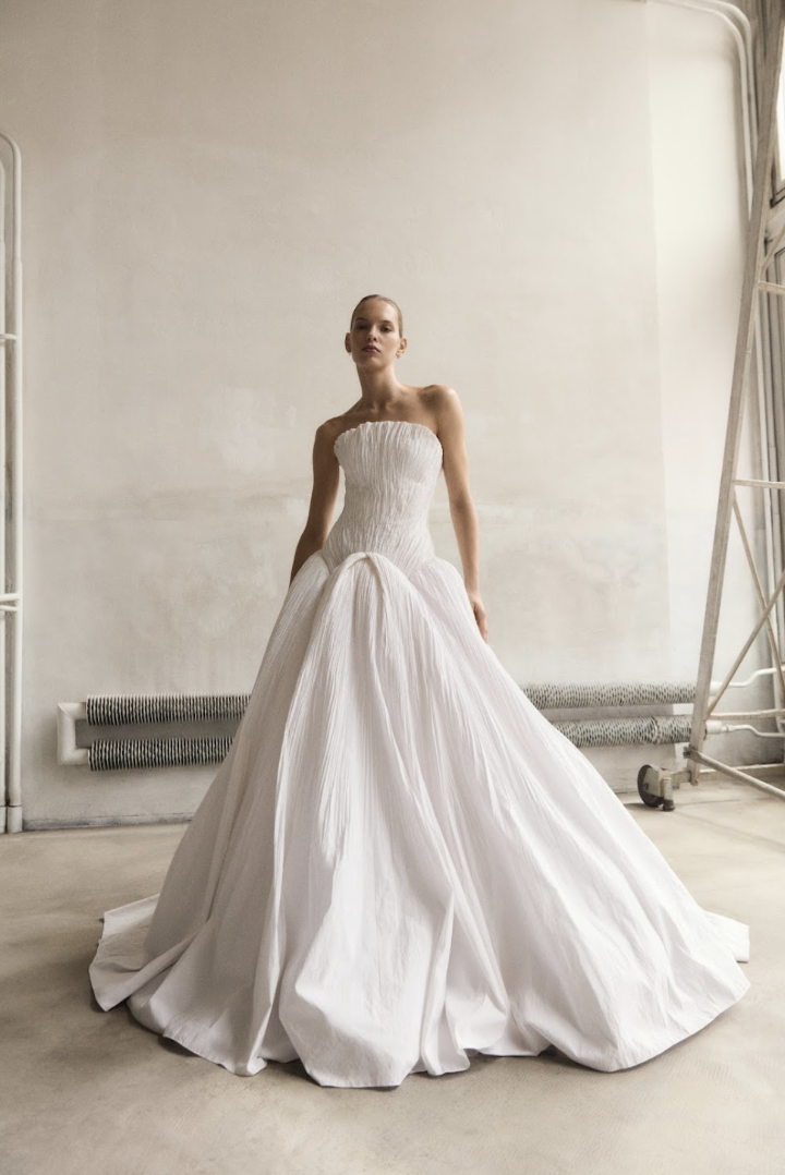 Marmar Halim at New York Bridal Market A woman in a strapless white wedding gown standing indoors against a plain wall.