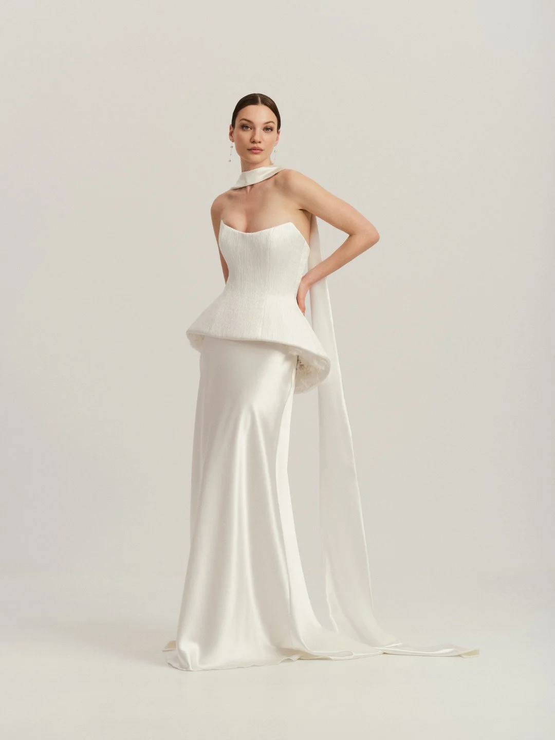 Divine Atelier at New York Bridal Market. Woman wearing an elegant off-white gown with a fitted bodice, long satin skirt, and matching cape against a plain background.