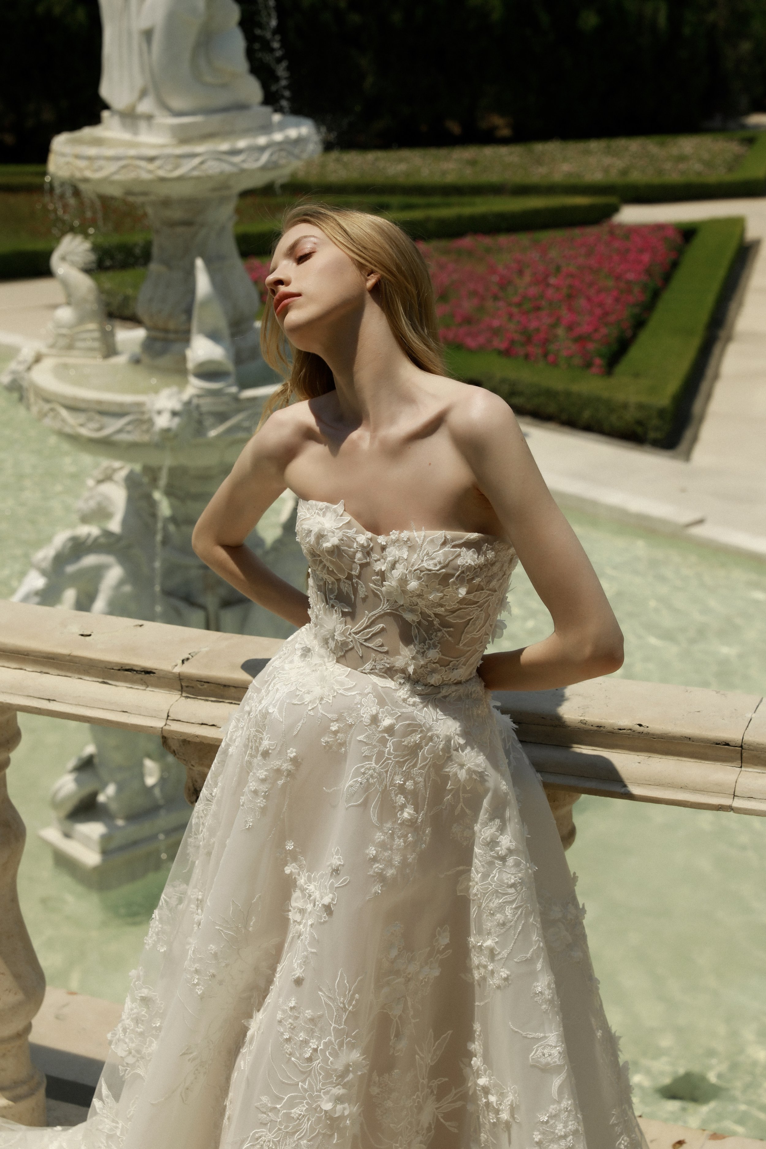 	
Guardiola at New York Bridal Market A woman in a strapless white wedding dress with floral embroidery standing in front of a fountain with a horse sculpture at a garden.