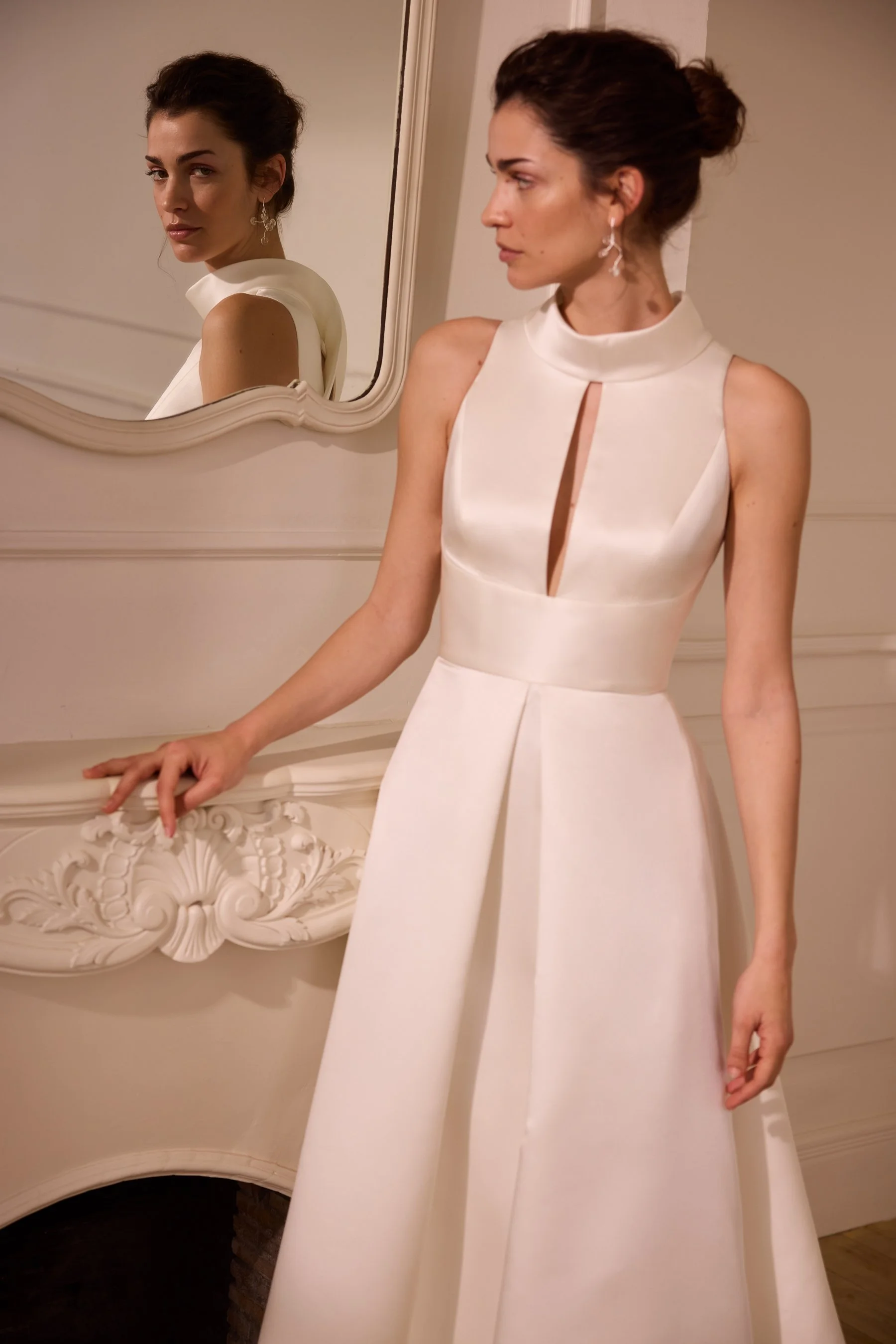 A woman in a white elegant dress standing in front of a mirror. Her reflection shows her face and upper body. The dress has a high neckline with a keyhole cutout. She is wearing drop earrings and has her hair styled in an updo. The background shows a