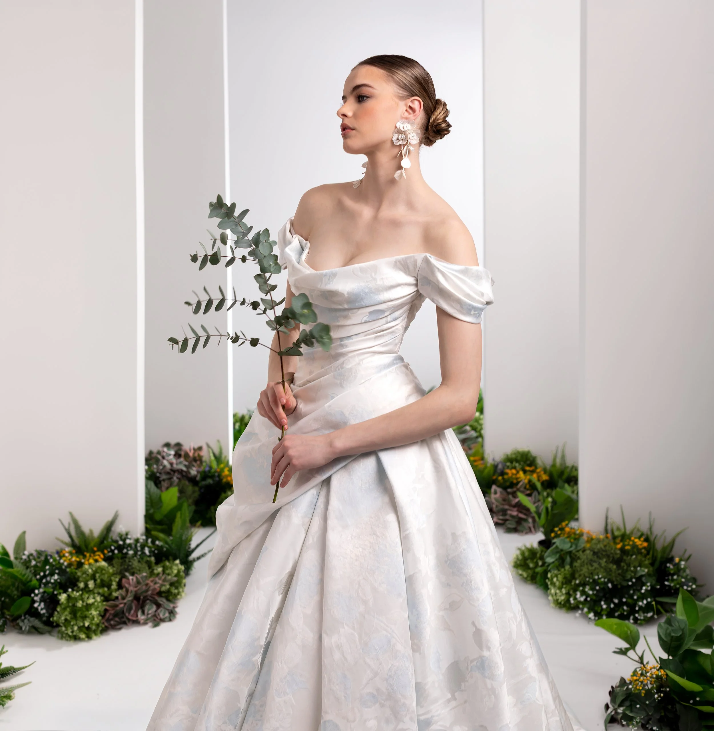 Valentini at New York Bridal Market. A woman with styled hair wearing a white off-shoulder gown and large white floral earrings, holding a eucalyptus branch, standing in front of a white backdrop with colorful potted plants at the bottom.