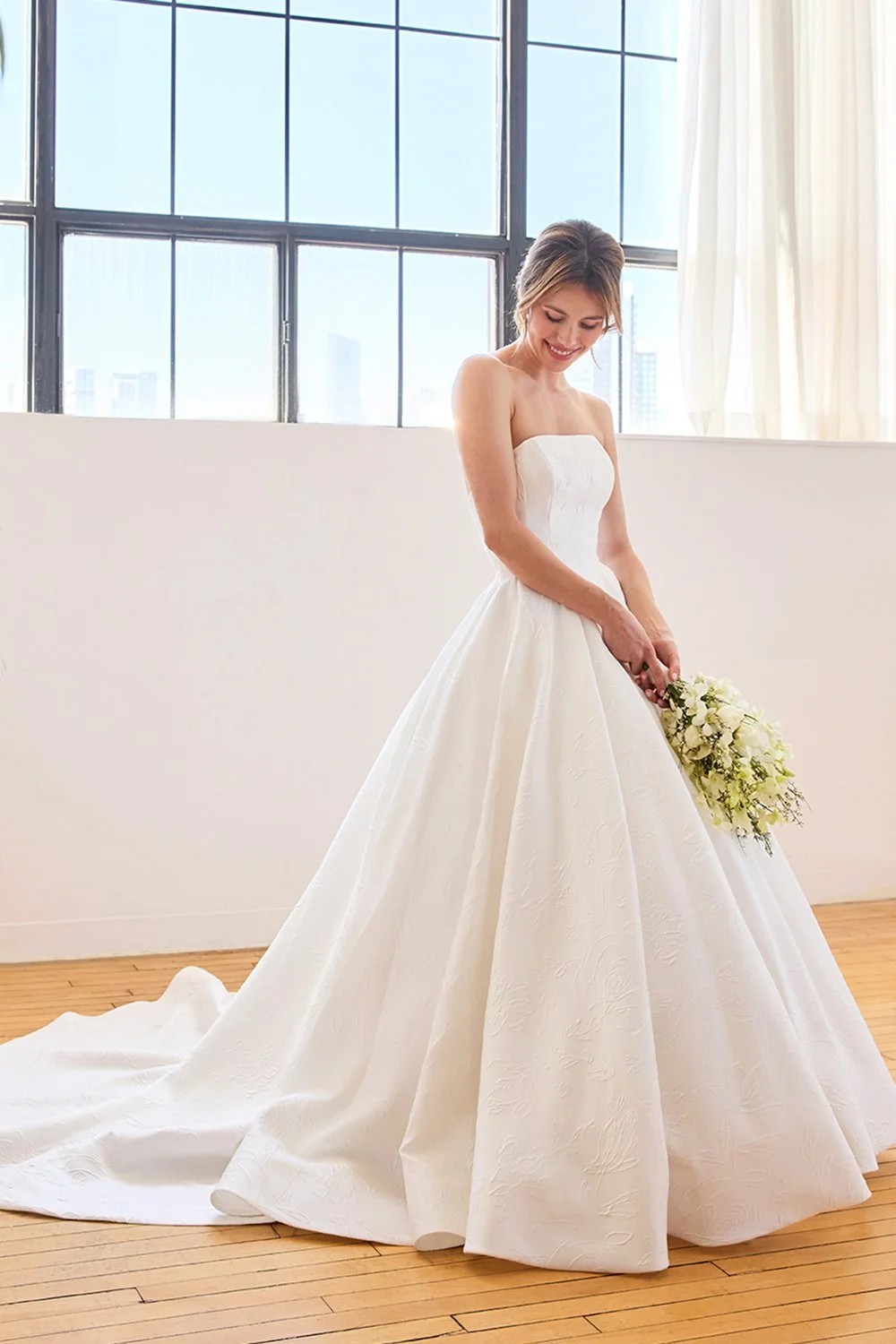 A bride in a strapless white wedding gown holding a bouquet of white flowers, standing indoors near large windows with a city skyline in the background.