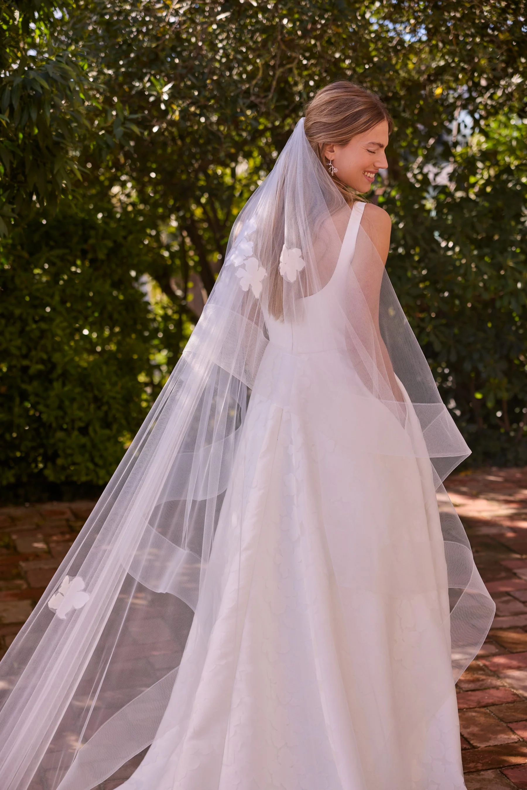 Jesus Peiro at New York Bridal Market.  A smiling woman in a white wedding dress and veil standing outdoors near greenery, looking to her right.