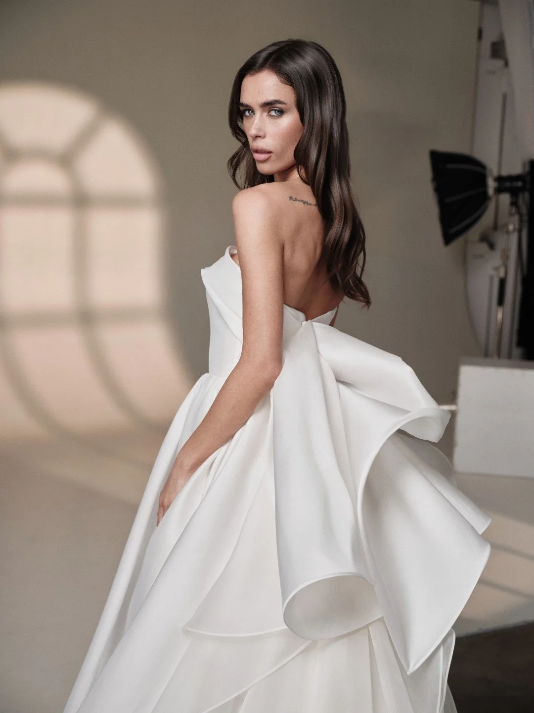Maria Pia at New York Bridal Market.  A woman in a white strapless gown with a large bow on the back, standing in a photography studio.