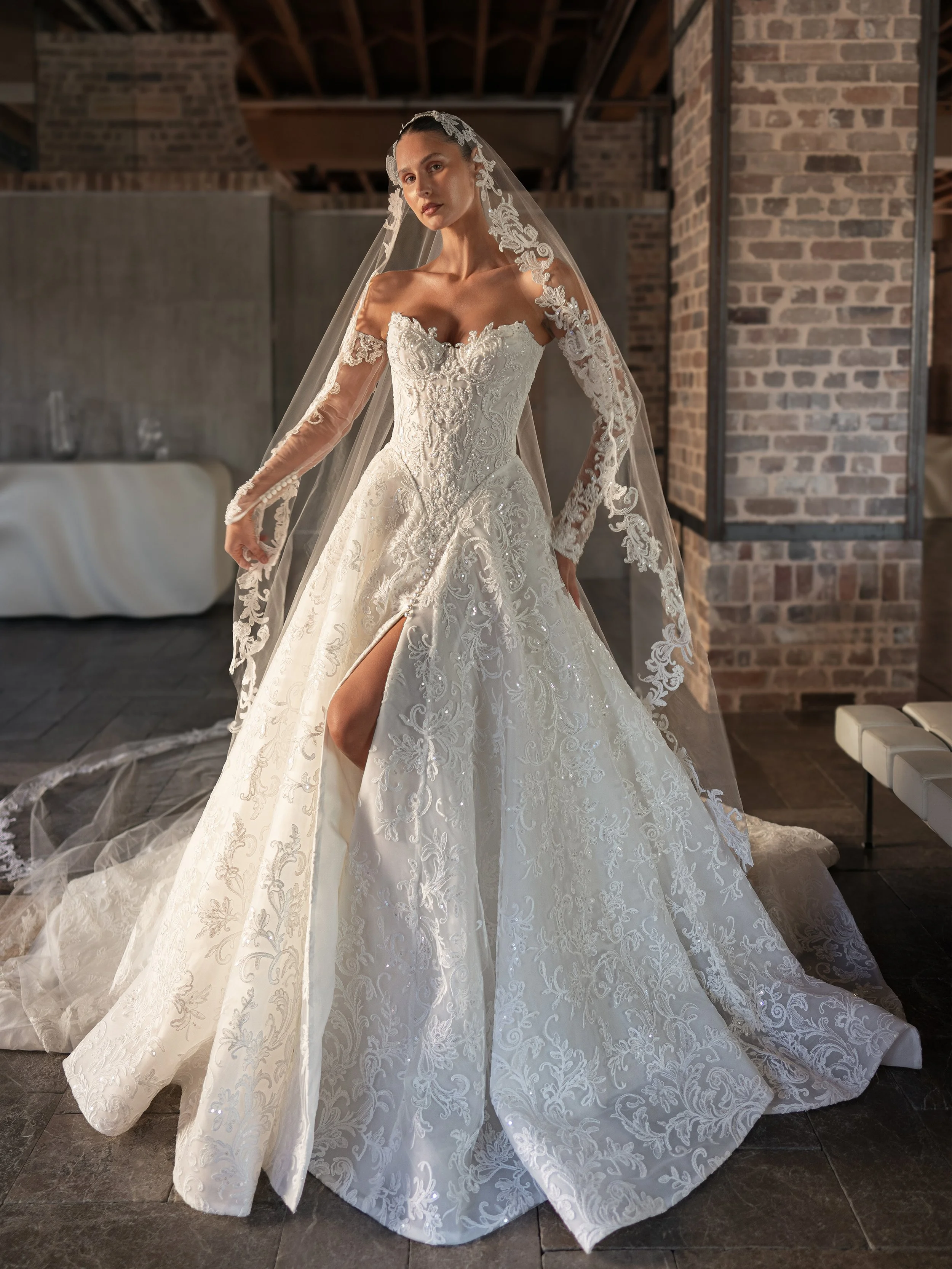 Blanche Bridal at New York Bridal Market. A woman wearing a wedding dress with lace details and a long train, standing indoors next to a brick wall and modern decor.