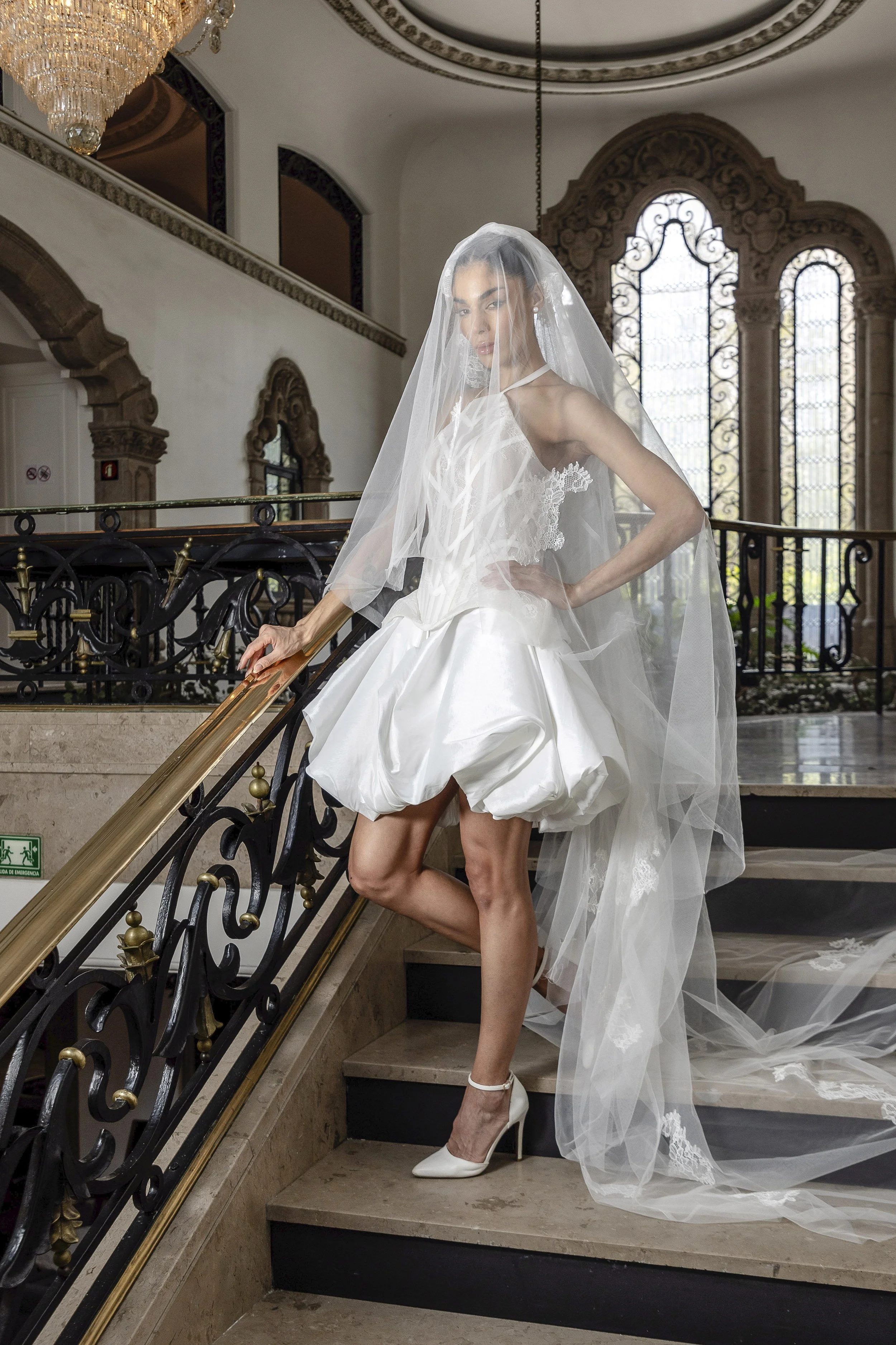 A woman in a wedding dress with a veil, high heels, and a tuxedo underneath, standing on a staircase inside a grand building with ornate windows and a chandelier.