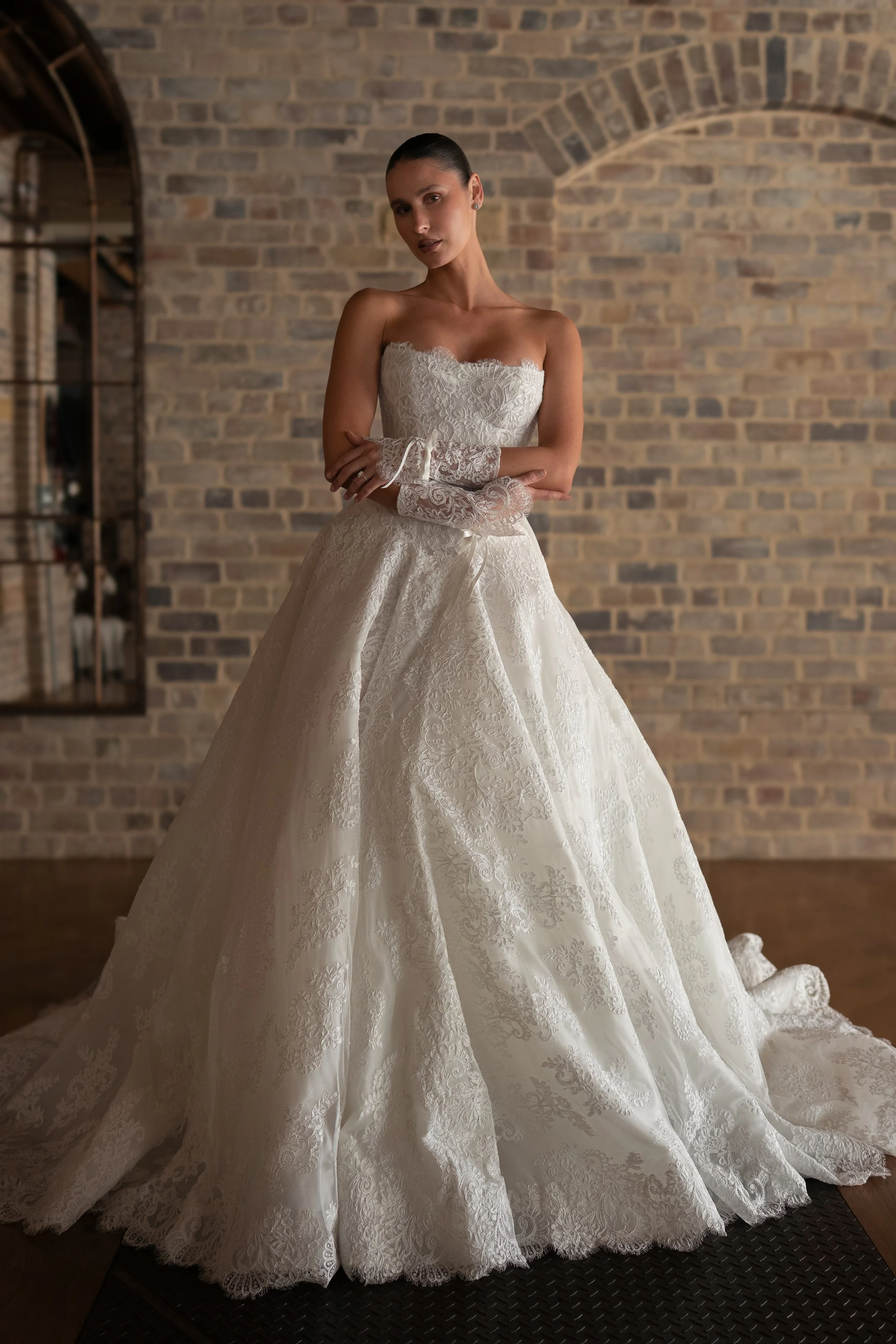 Blanche Bridal at New York Bridal Market  A woman in a white wedding gown with lace details, standing against a brick wall, with her arms crossed and a serious expression.