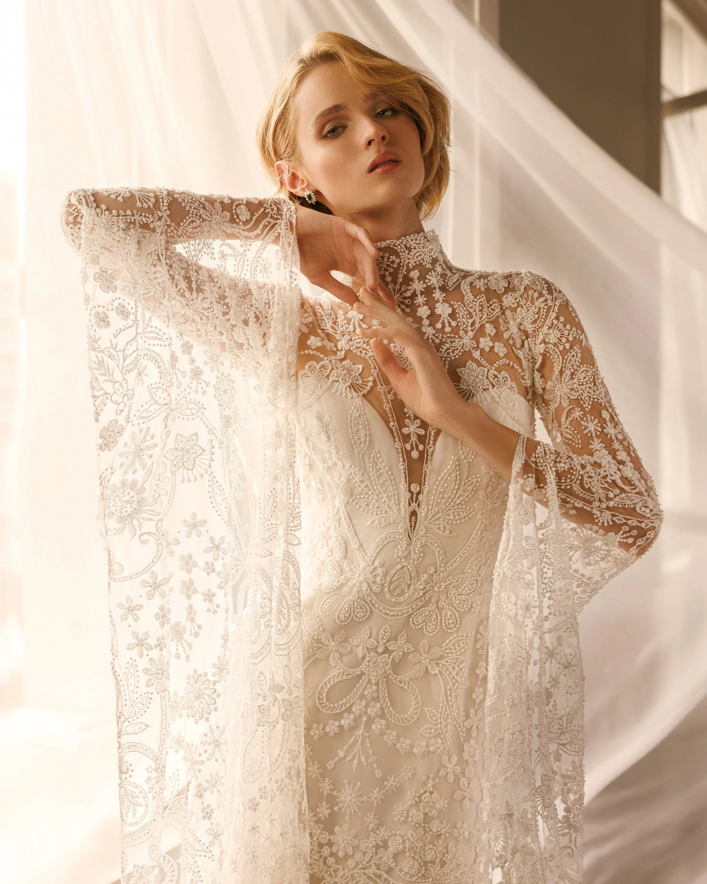 Gemy Maalouf at New York Bridal Makrket A woman with short blonde hair, wearing a lace dress and floral earrings, poses indoors with a neutral expression, looking slightly up and to the side.
