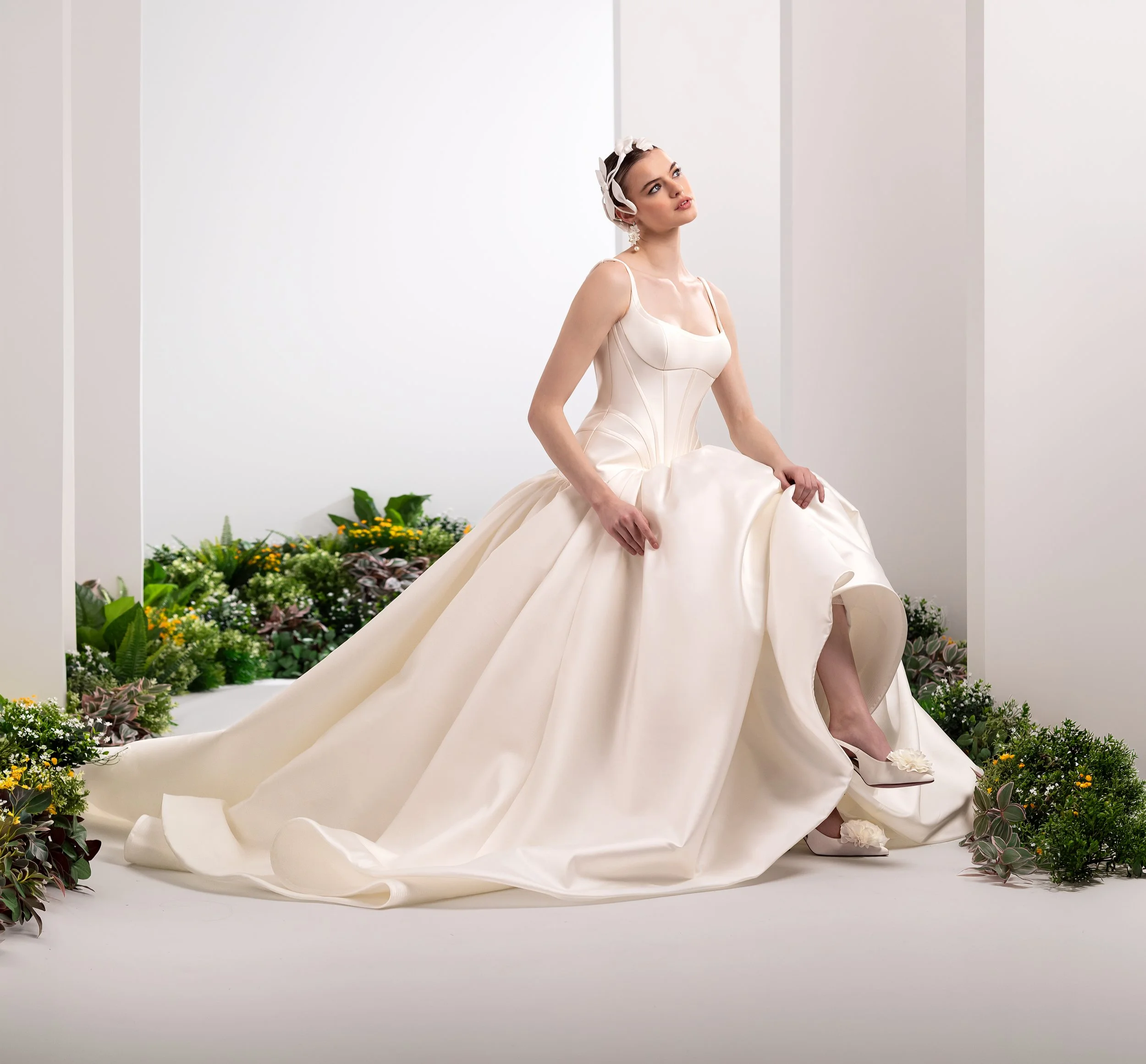 Valentini Spose at New York Bridal Market.   A woman in an elegant ivory gown with a full skirt sitting on her heels, surrounded by flowers and greenery on a white background, possibly at a fashion or wedding event.