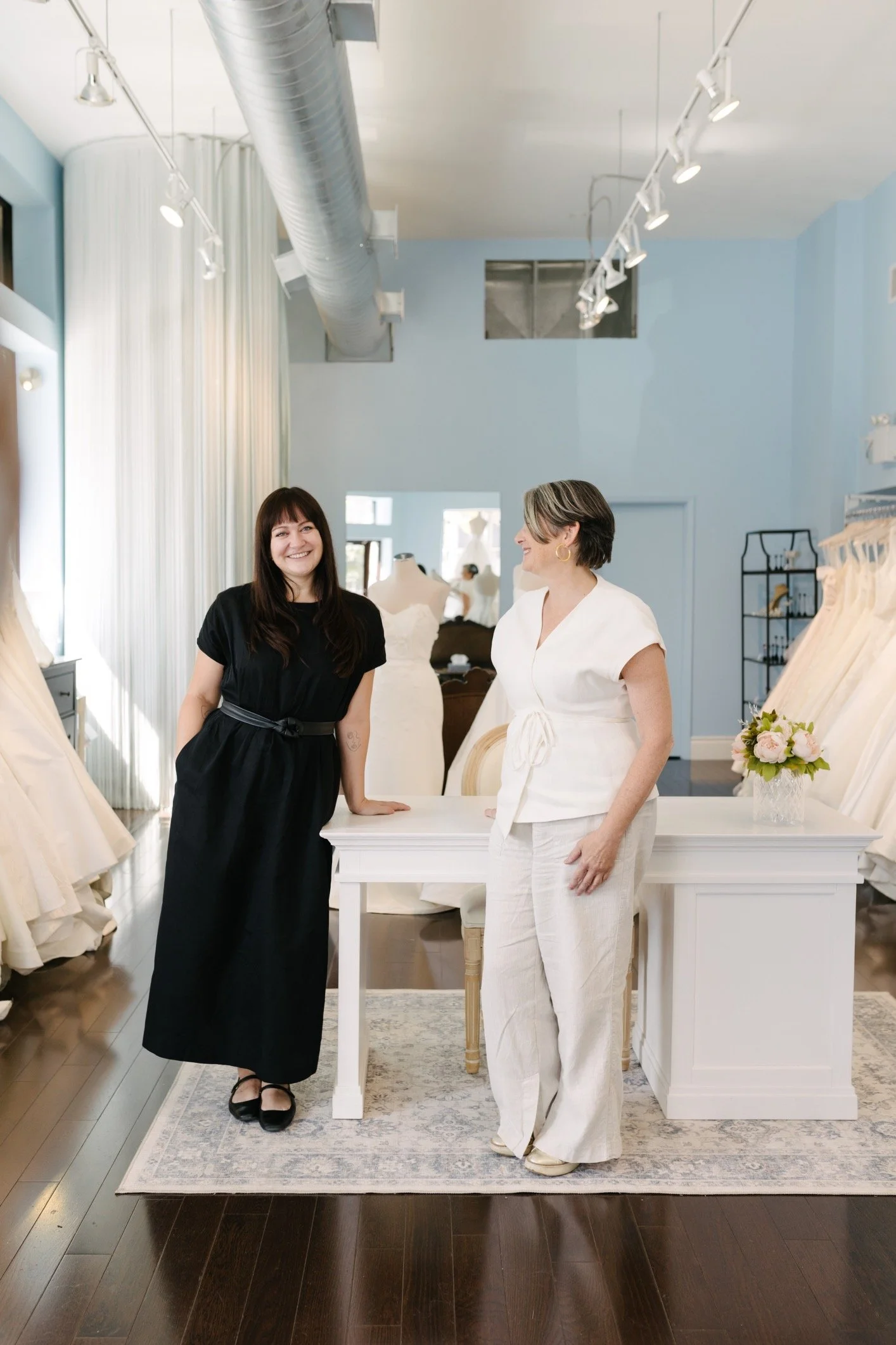 Lea-Ann Belter at New York Bridal Market. Two women standing in a boutique or studio, surrounded by dresses and mannequins, inside a brightly lit space with light blue walls and wooden floors.