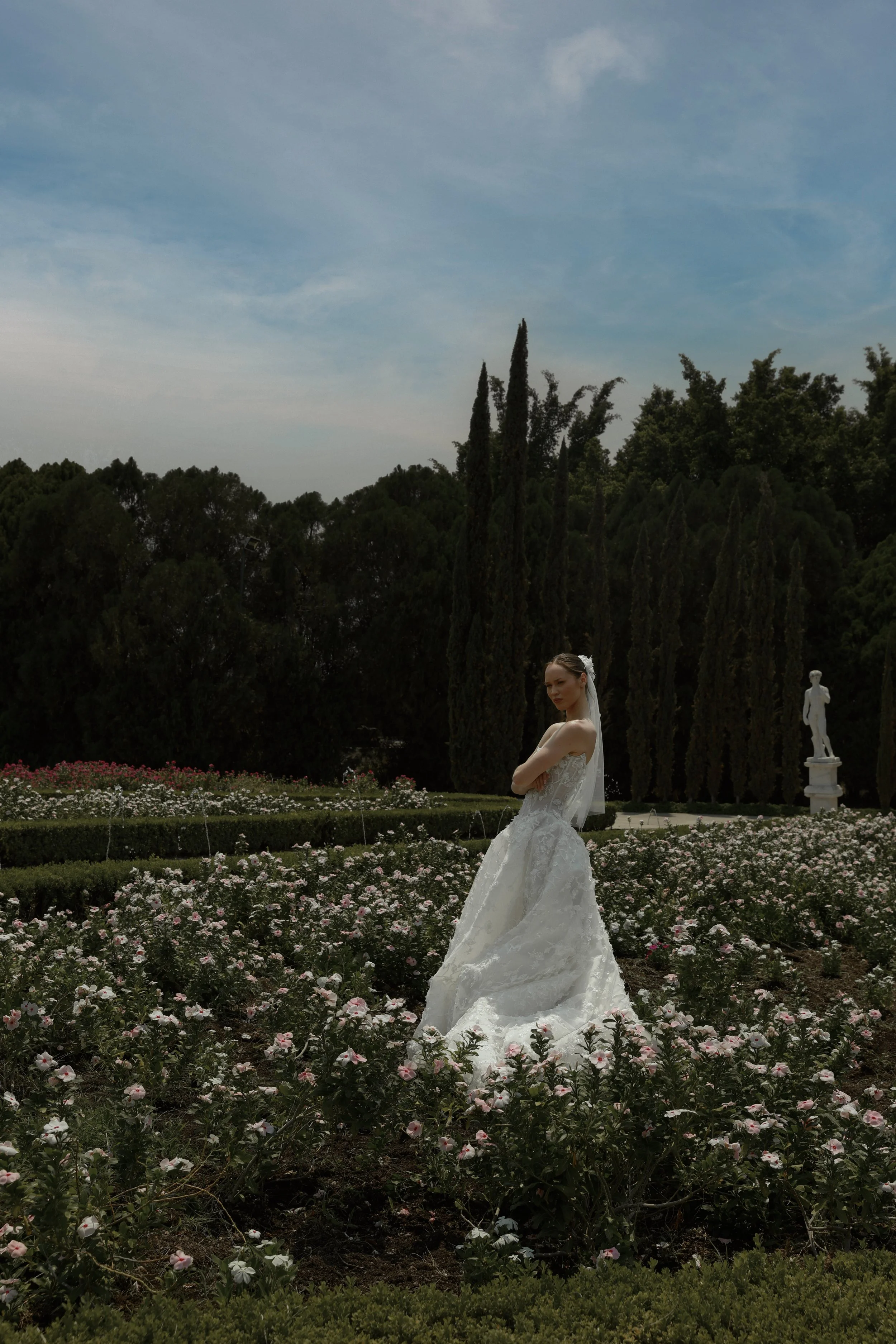 	
Guardiola at New York Bridal Market A bride in a white wedding gown with a veil standing in a garden filled with pink and white flowers, with tall trees and blue sky in the background.