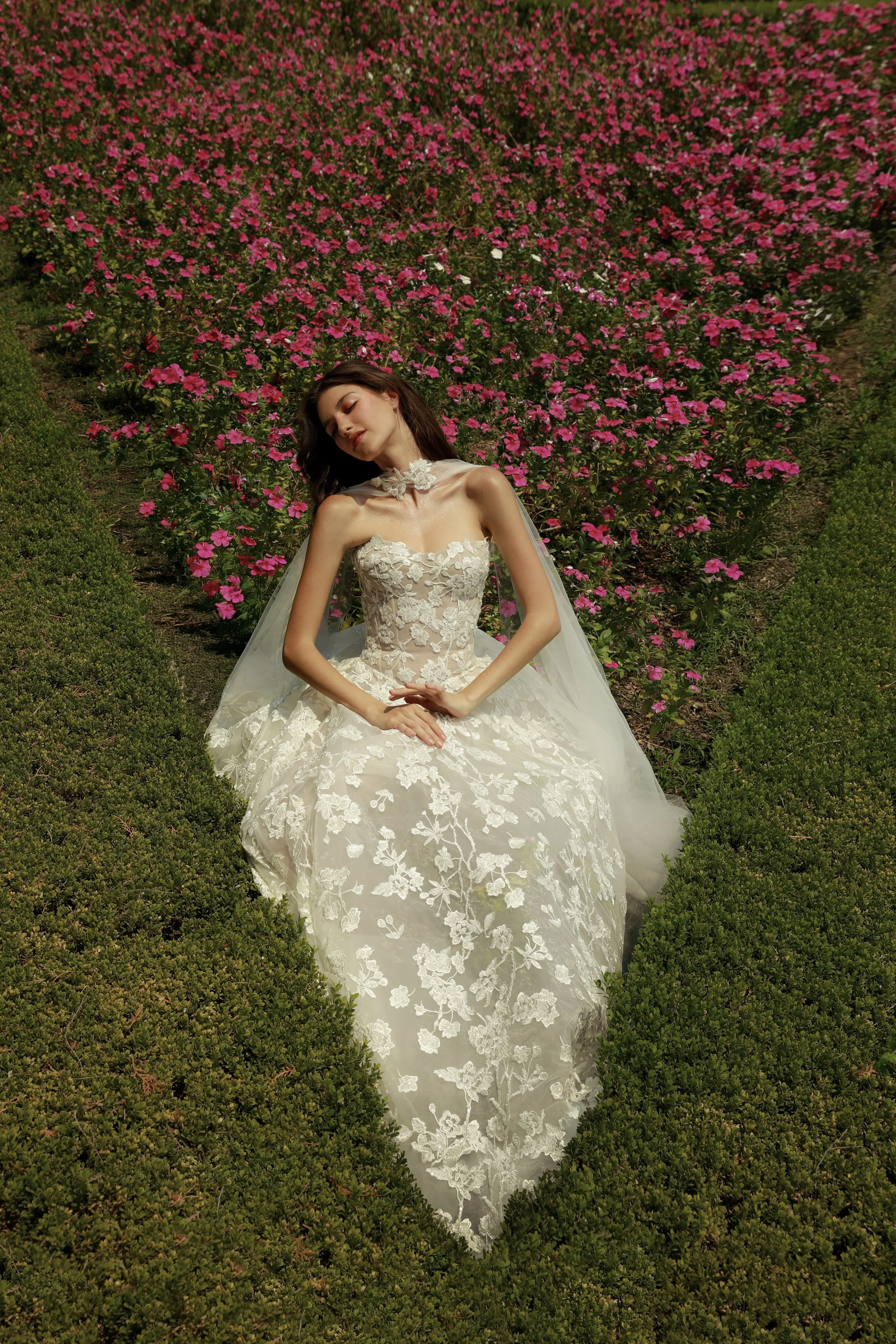 Guardiola Bridal at New York Bridal Market. A woman in a white wedding dress sitting with closed eyes in a field of pink flowers.