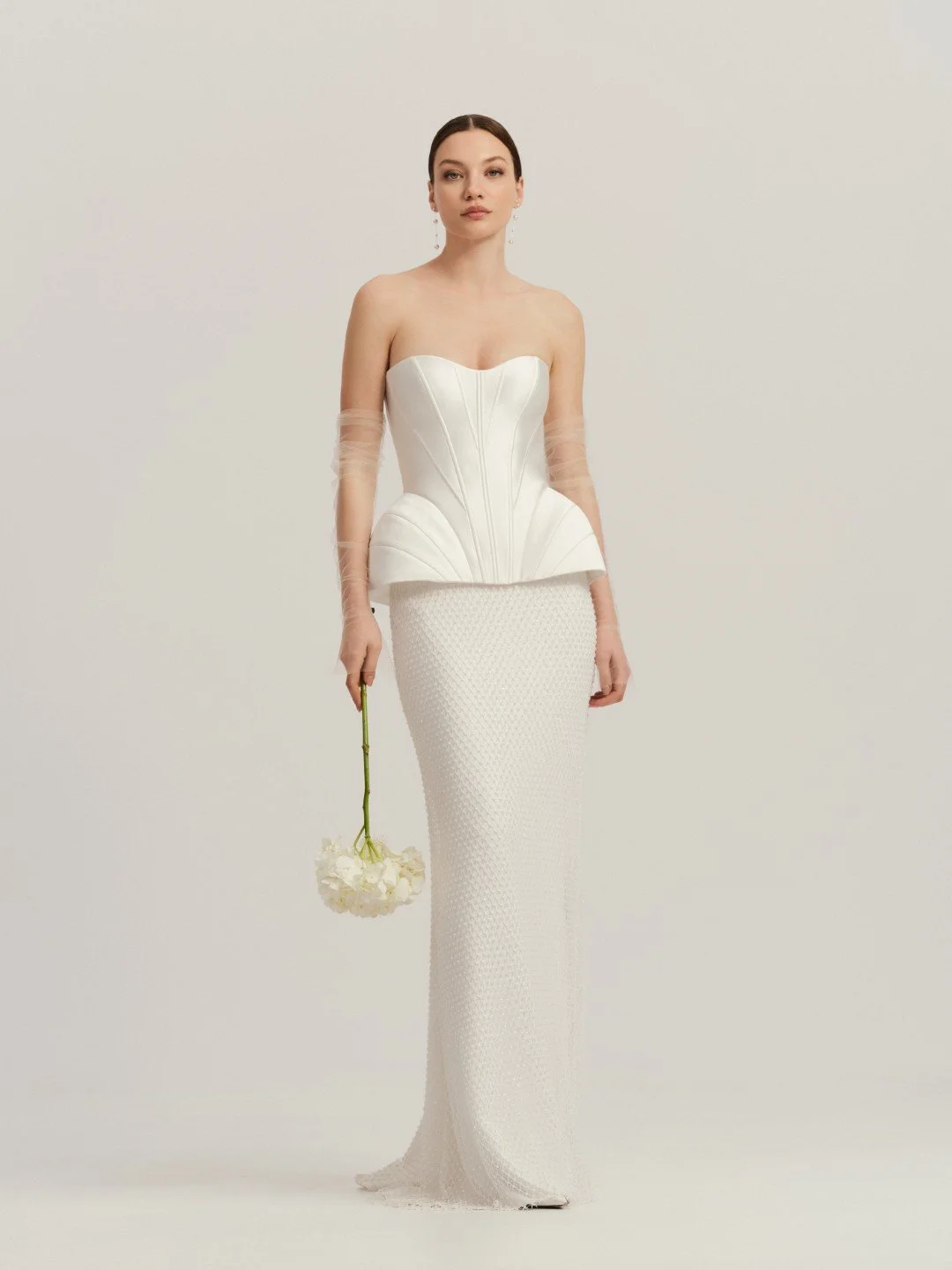Divine Atalier at New York Bridal Market. A woman in a white strapless dress with a fitted bodice and textured skirt holding a white flower in her right hand against a plain background