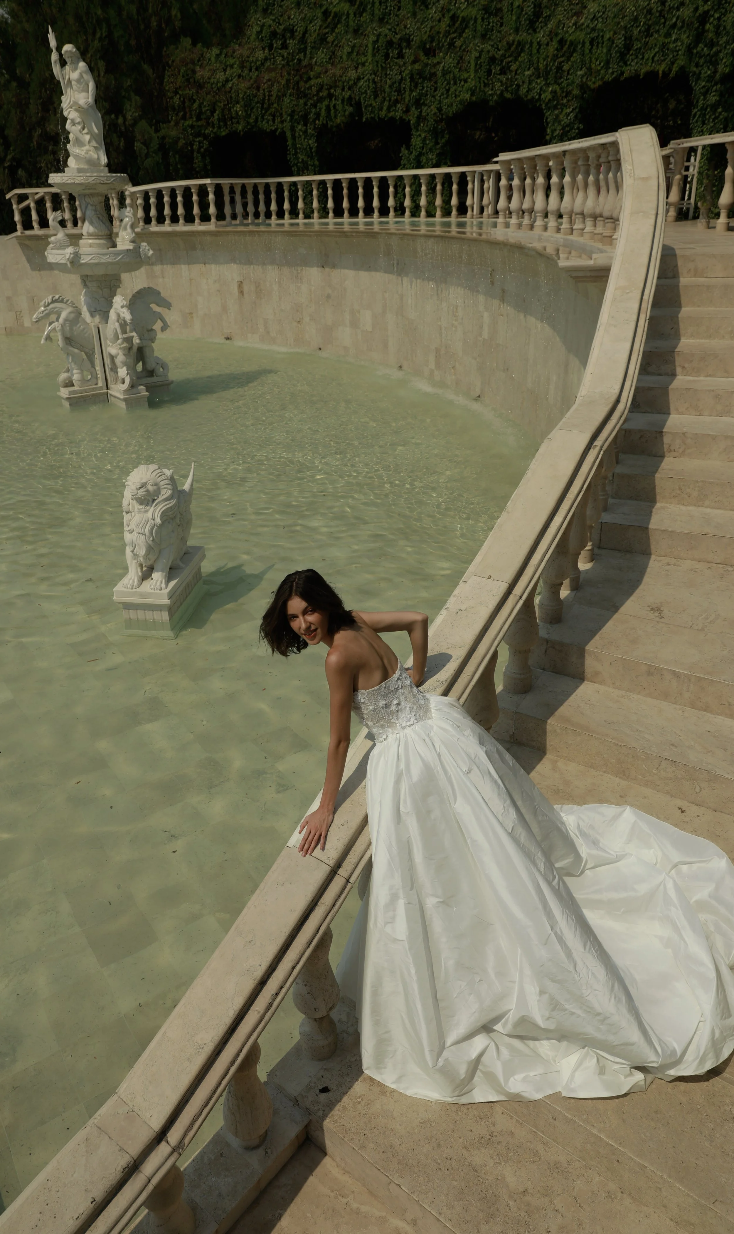 	
Guardiola at New York Bridal Market A woman in a white wedding dress leaning on a railing near a fountain with statues, on a stone staircase outside.