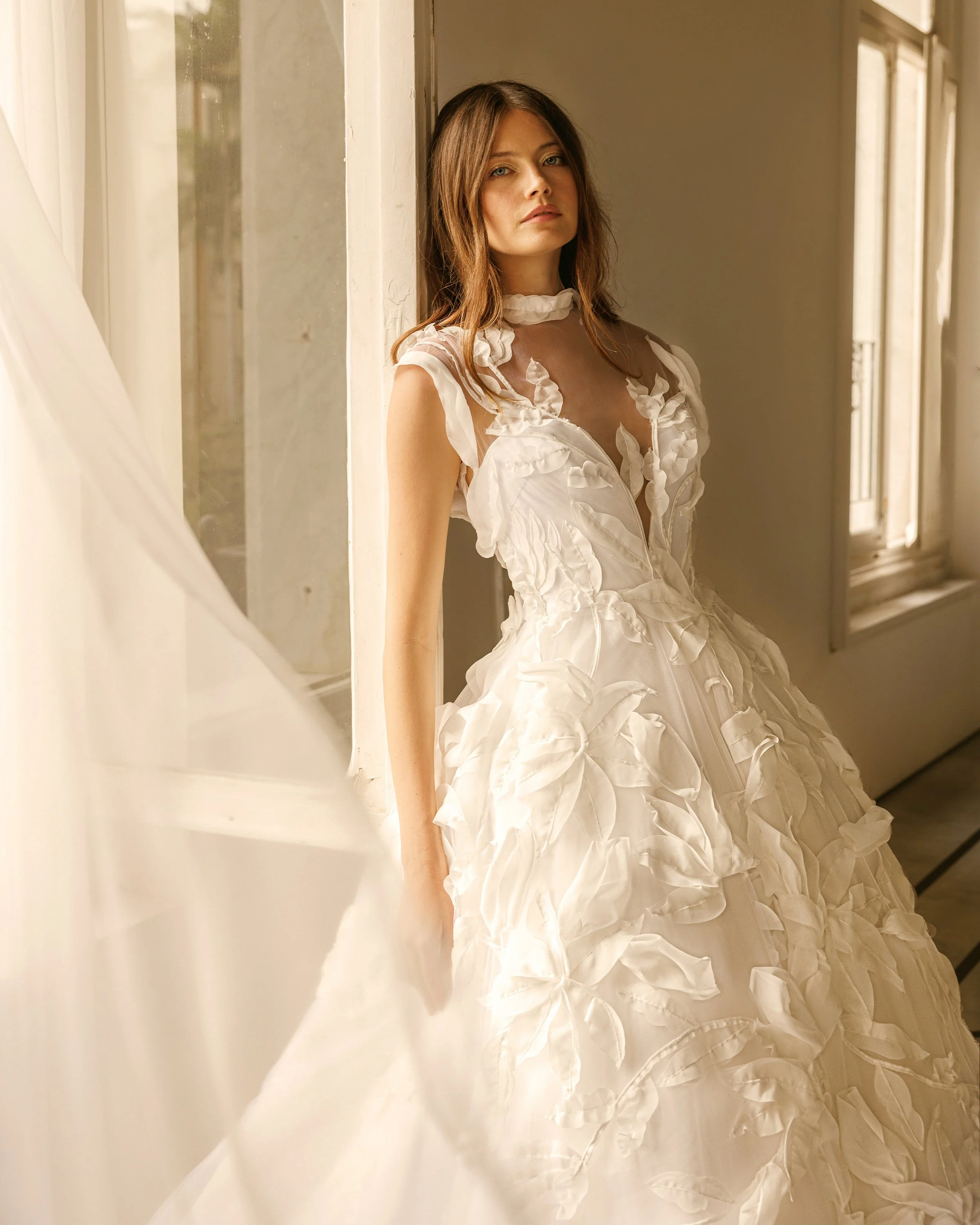 Gemy Maalouf at New York Bridal Makrket. A woman with long brown hair wearing a white dress with ruffled details, leaning against a window frame in a softly lit room.