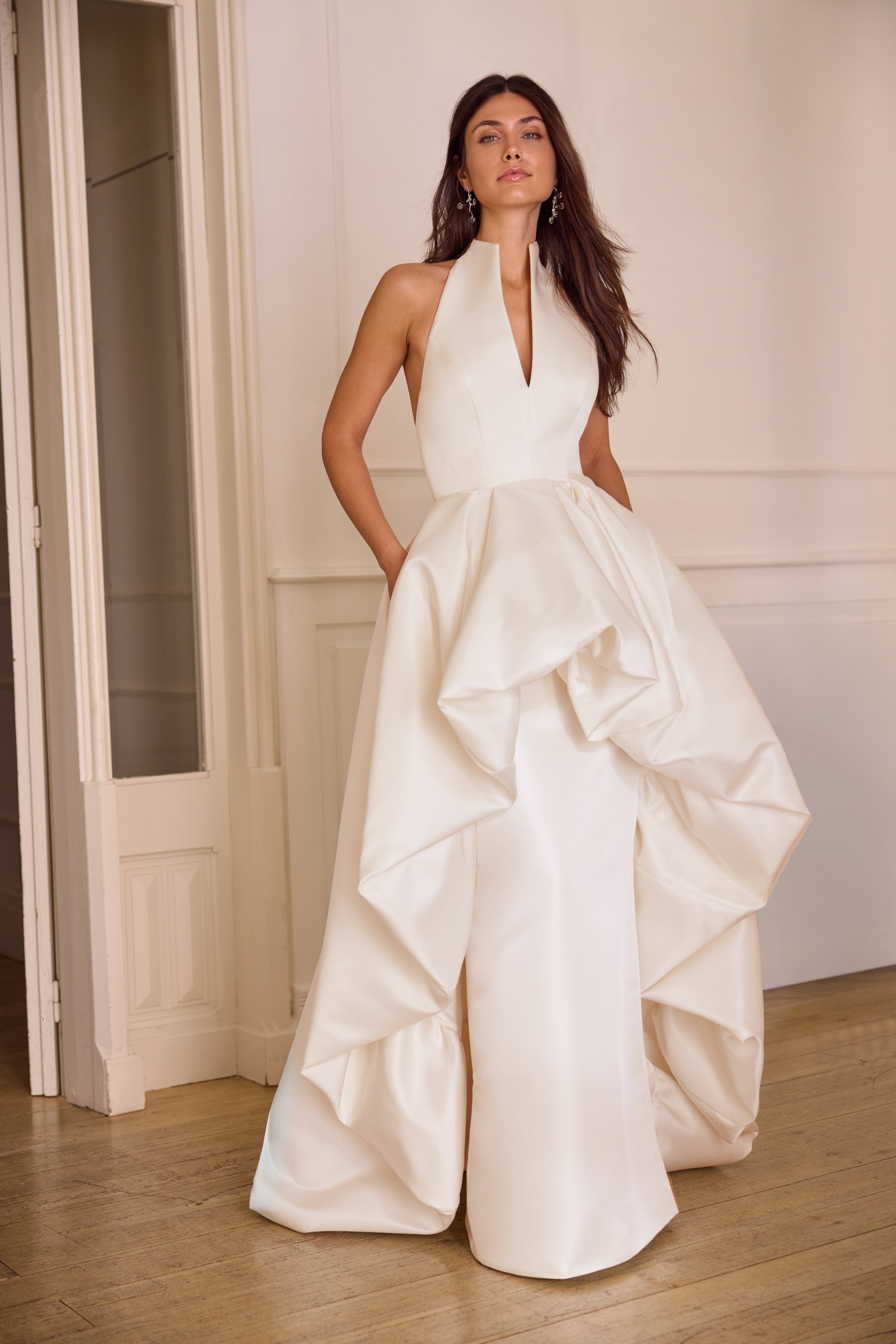 Jesus Peiro at New York Bridal Market.  Woman in a white formal gown standing in a room with wood flooring, posing with her hands in her pockets and looking at the camera.