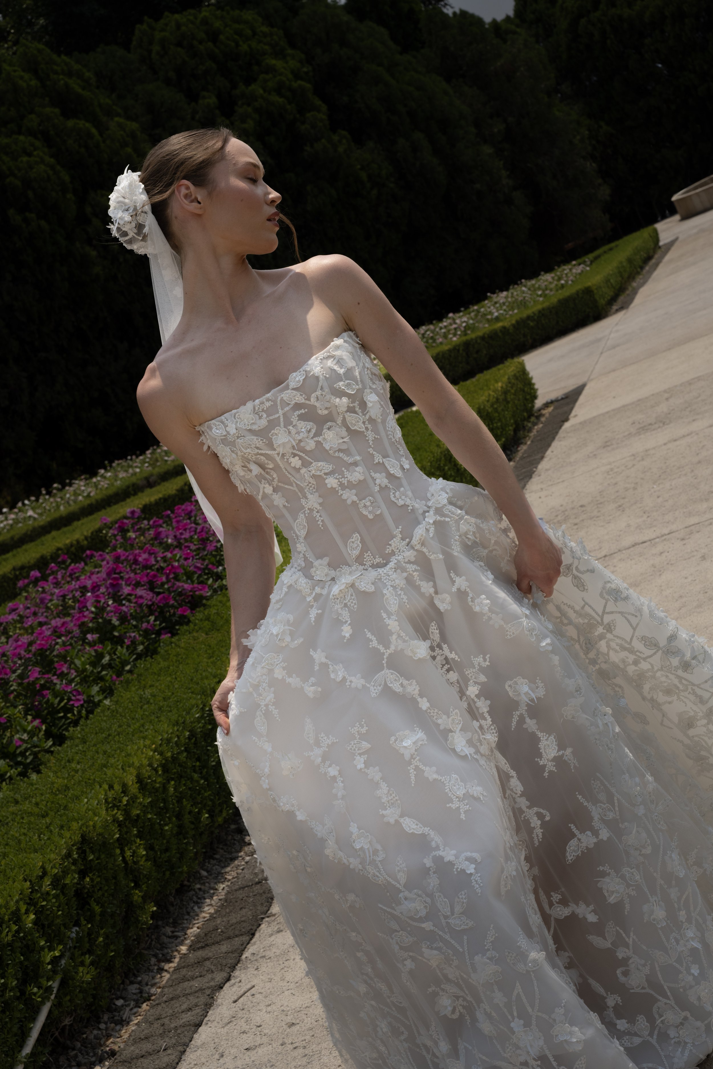 A woman in a white lace wedding gown with floral embroidery, standing outdoors near flower beds and greenery, with her eyes closed and head slightly turned.