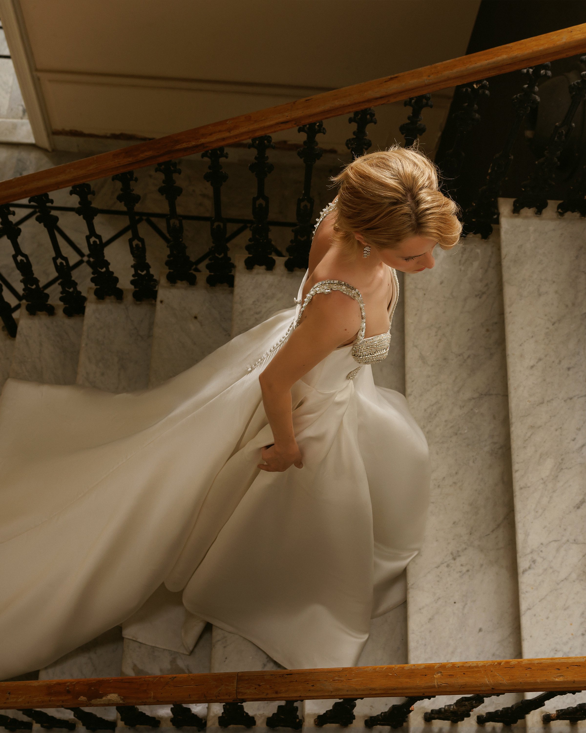 Gemy Maalouf at New York Bridal Makrket A bride in a white wedding gown with embellishments on the shoulders, sitting on marble stairs at the bottom of a staircase with metal railings and a wooden handrail.
