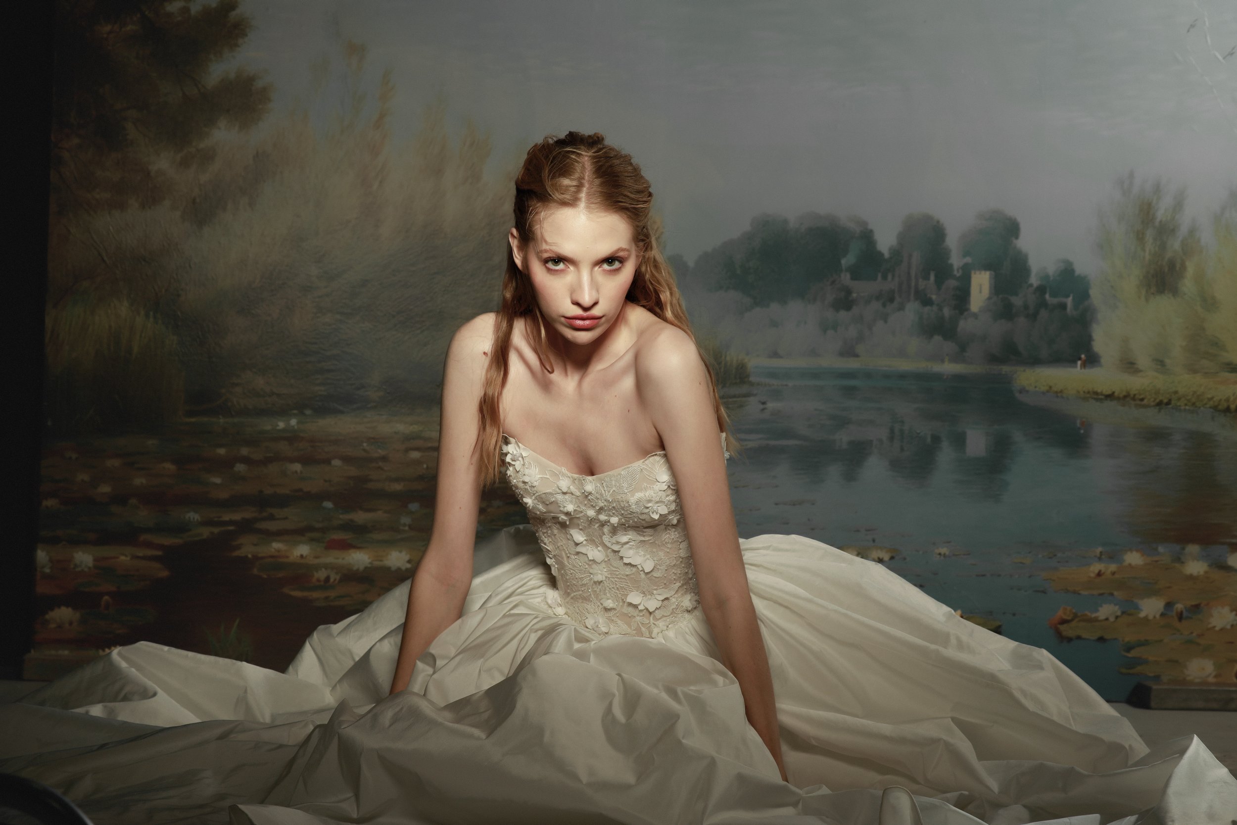 	
Guardiola at New York Bridal Market A woman in an elegant, strapless lace dress sitting on the ground in front of a painted landscape backdrop featuring a river, trees, and distant houses.