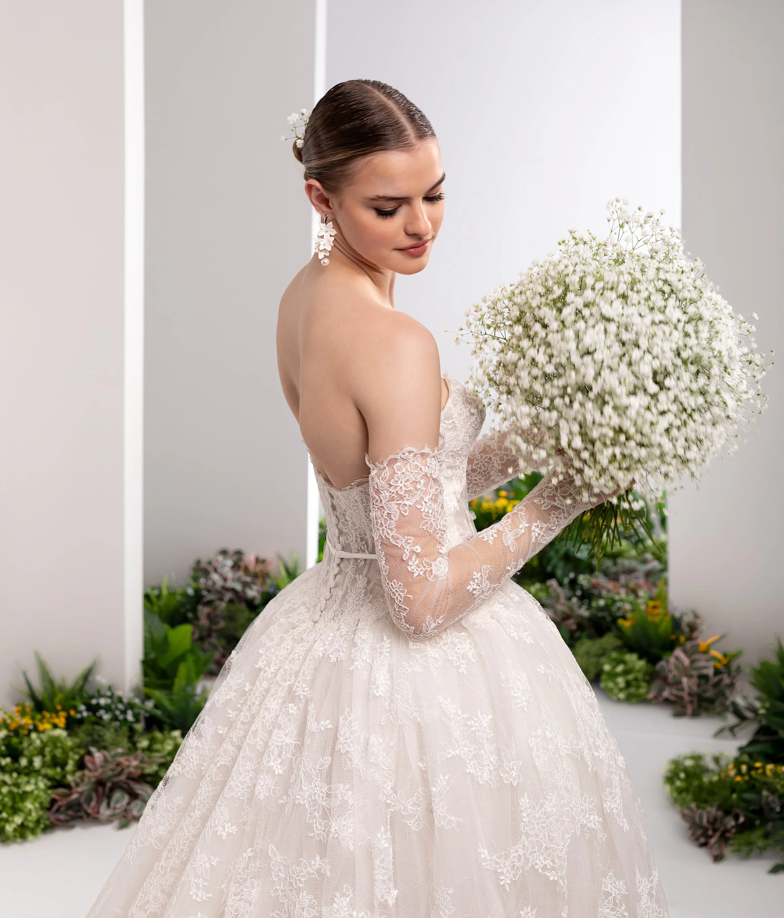 Valentini Spose at New York Bridal Market.  A bride with braided hair styled in a low bun decorated with baby's breath flowers, wearing a lace wedding gown with long sleeves, posed in front of a neutral background with floral arrangements.