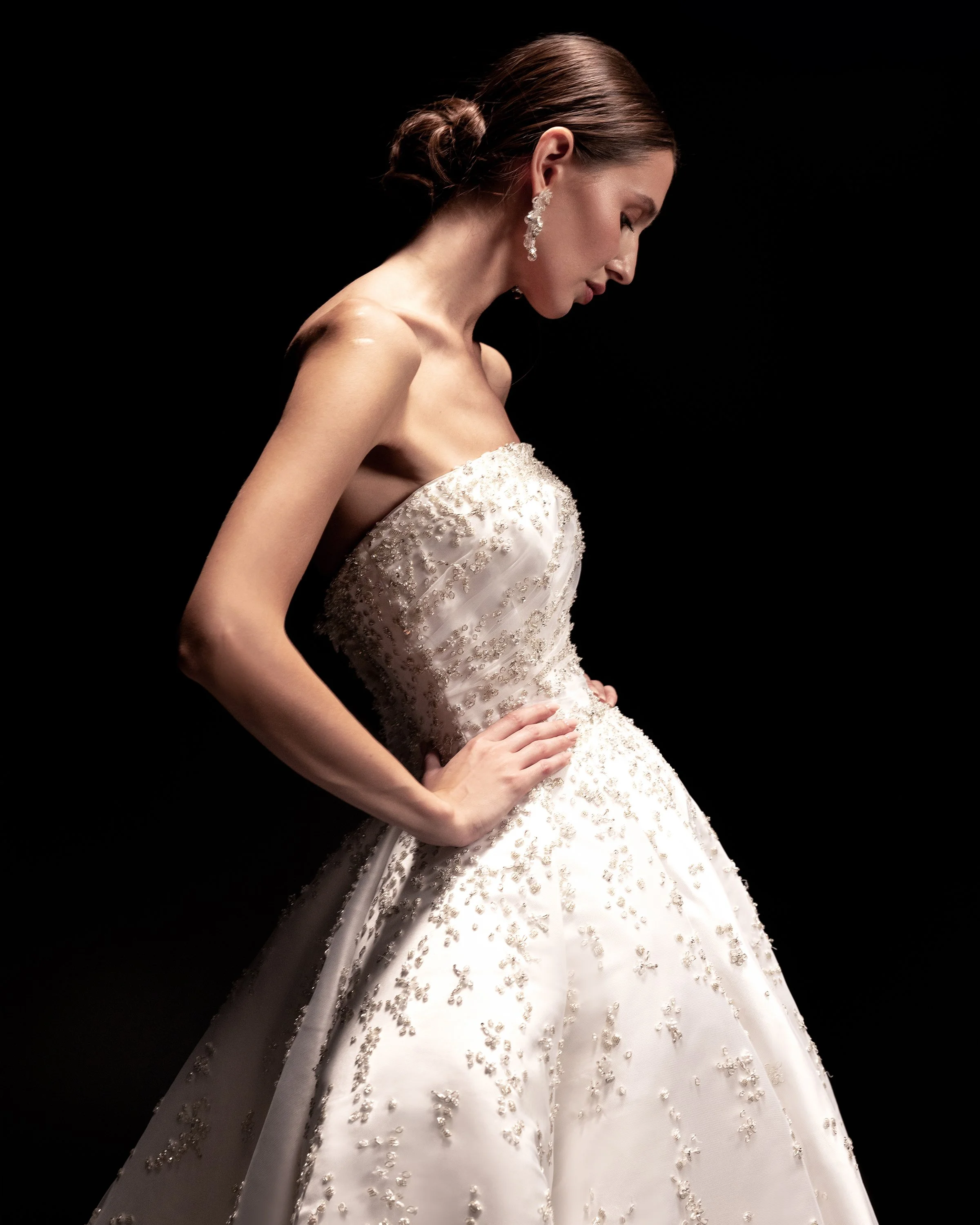 Gemy Maalouf at New York Bridal Makrket A woman in an elegant white wedding dress with intricate beaded embroidery, her hand on her hip, against a black background.