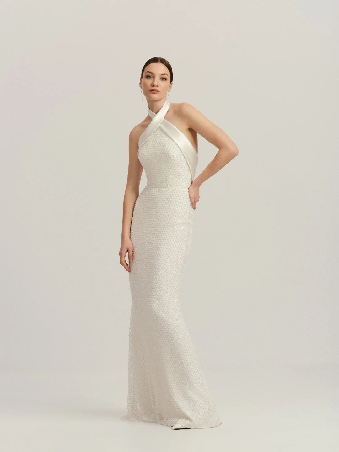 Divine Atelier at New York Bridal Market.  A woman wearing an elegant white evening gown with a halter neckline and textured fabric, standing against a cream background.