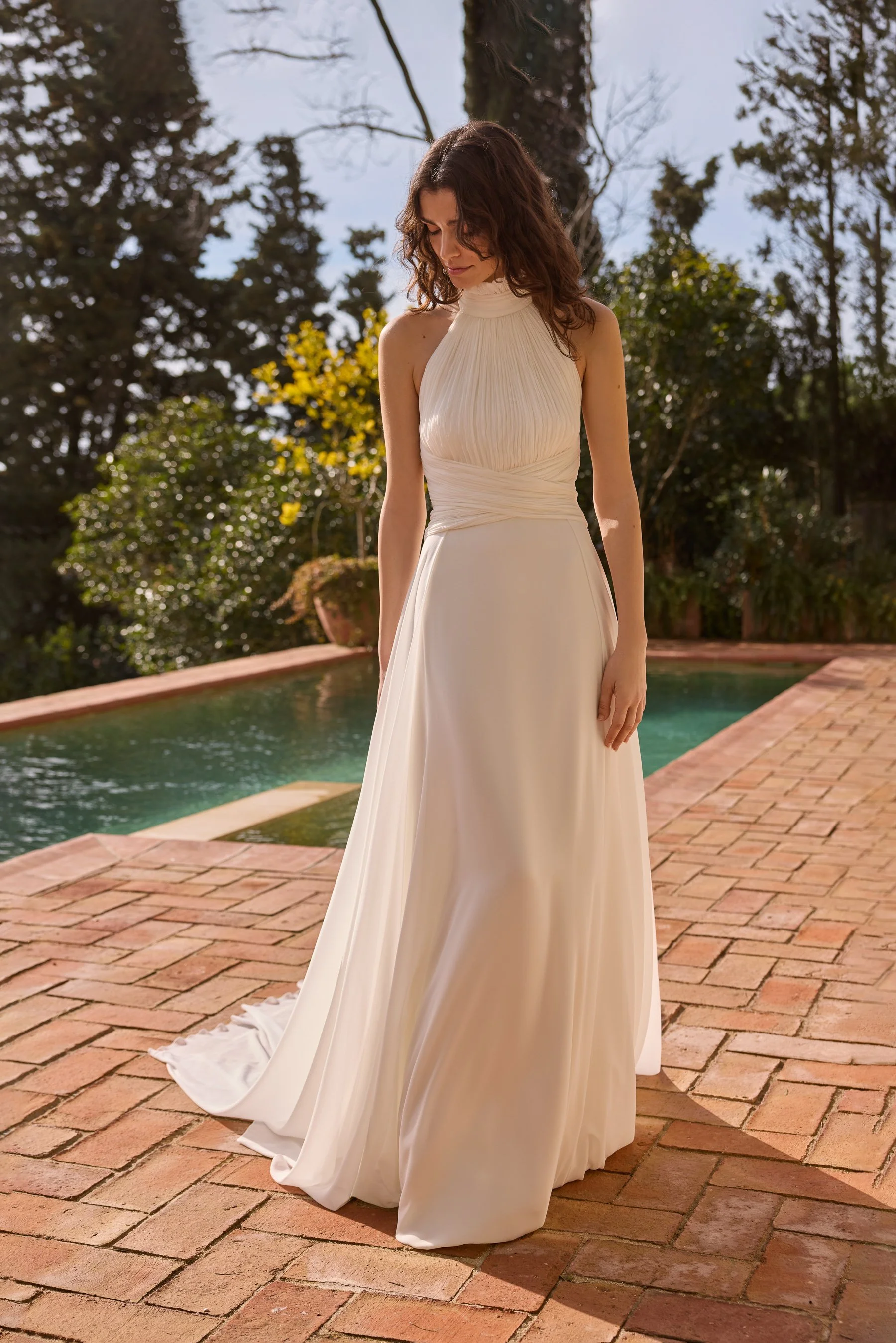 Jesus Peiro at New York Bridal Market.  A woman in a cream-colored, high-neck gown with pleats is standing outdoors on a brick patio near a pool, surrounded by trees with yellow and green leaves, under a partly cloudy sky.