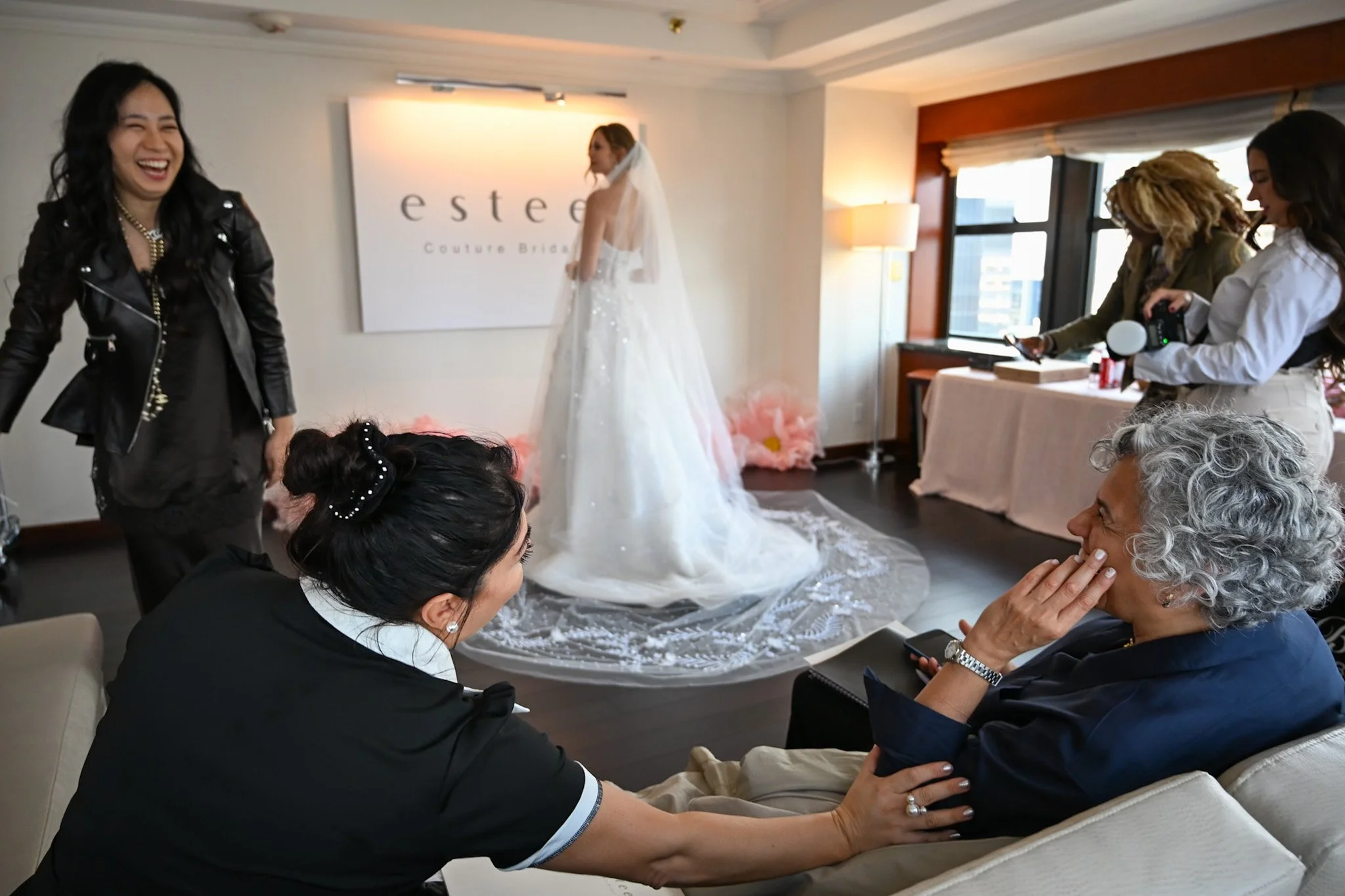 Women at a wedding dress fitting session, one in a bridal gown, others assisting and taking photos, in a brightly lit room with a large window.