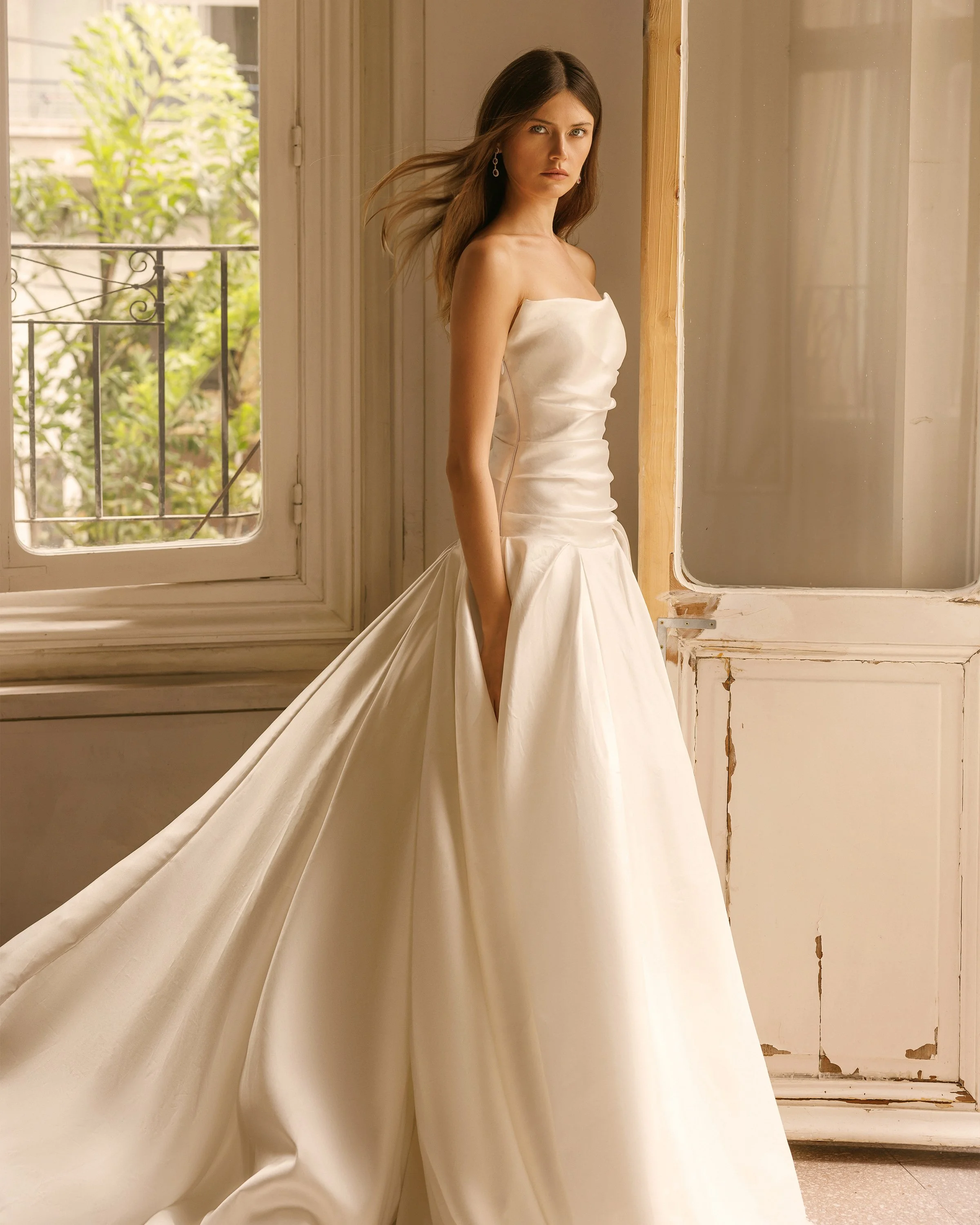 A woman with long brown hair standing indoors in a strapless white satin wedding gown with a full skirt and a slit, looking directly at the camera with a serious expression. She is wearing earrings, and there are windows behind her showing a tree out