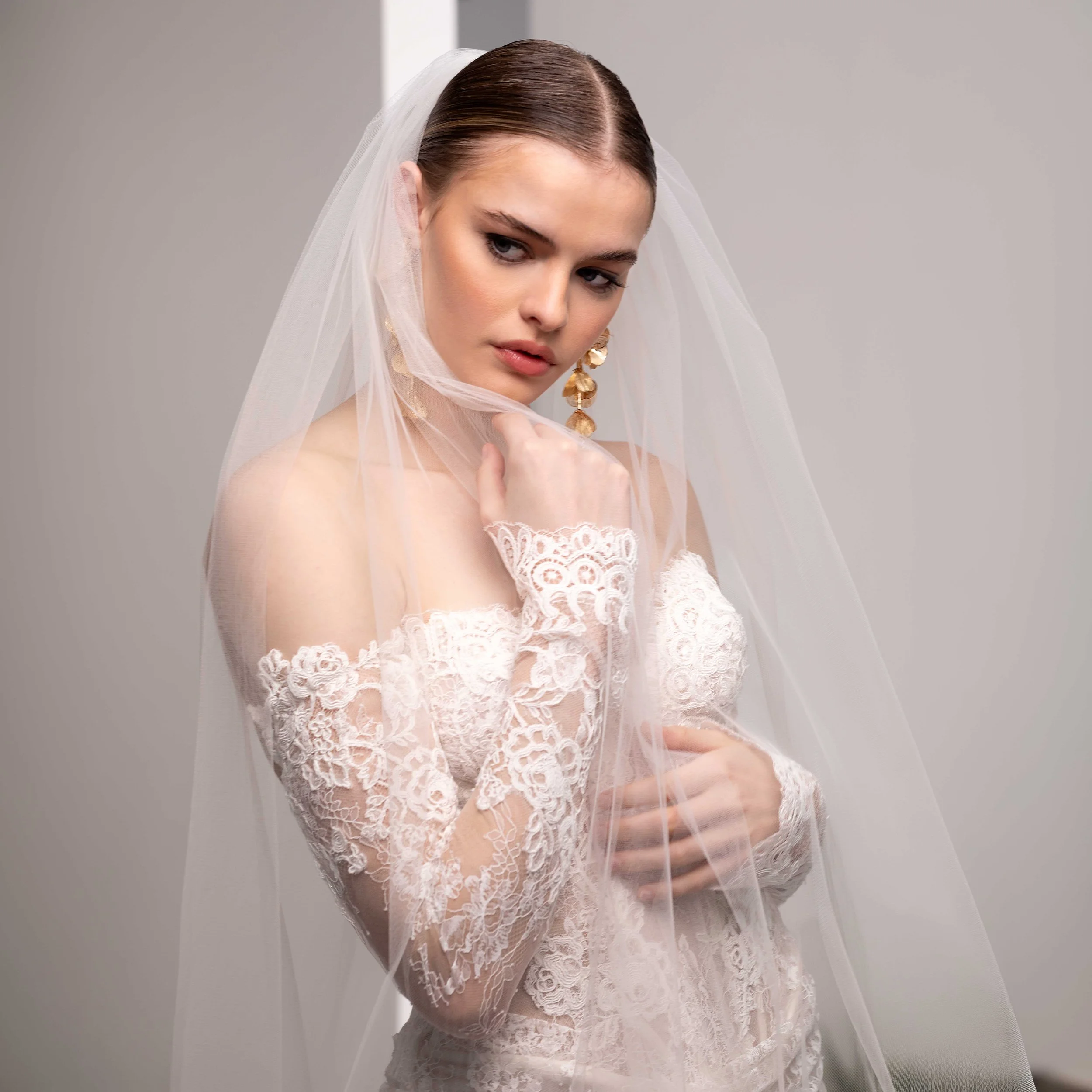 Valentini Spose at New York Bridal Market.   A portrait of a woman in a white lace dress and a white veil, looking down with her hand near her face, against a plain gray background.