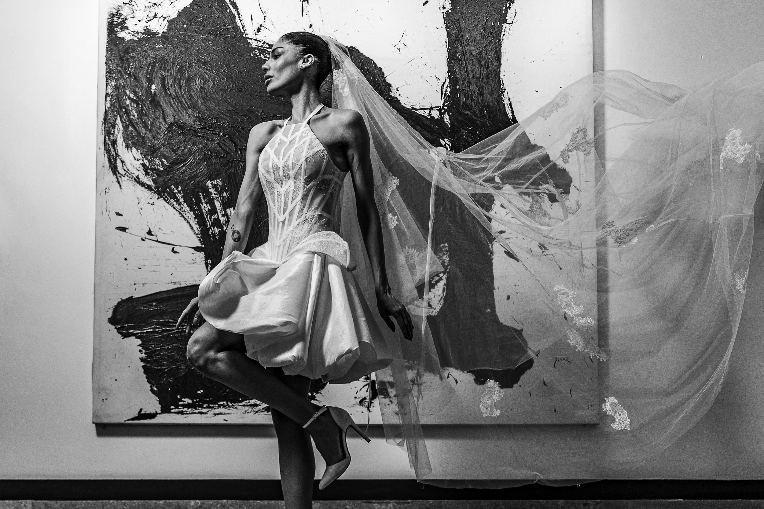 A woman in a wedding dress standing in front of an abstract painting, with her head turned to the side and her right leg crossed in front of her left leg.