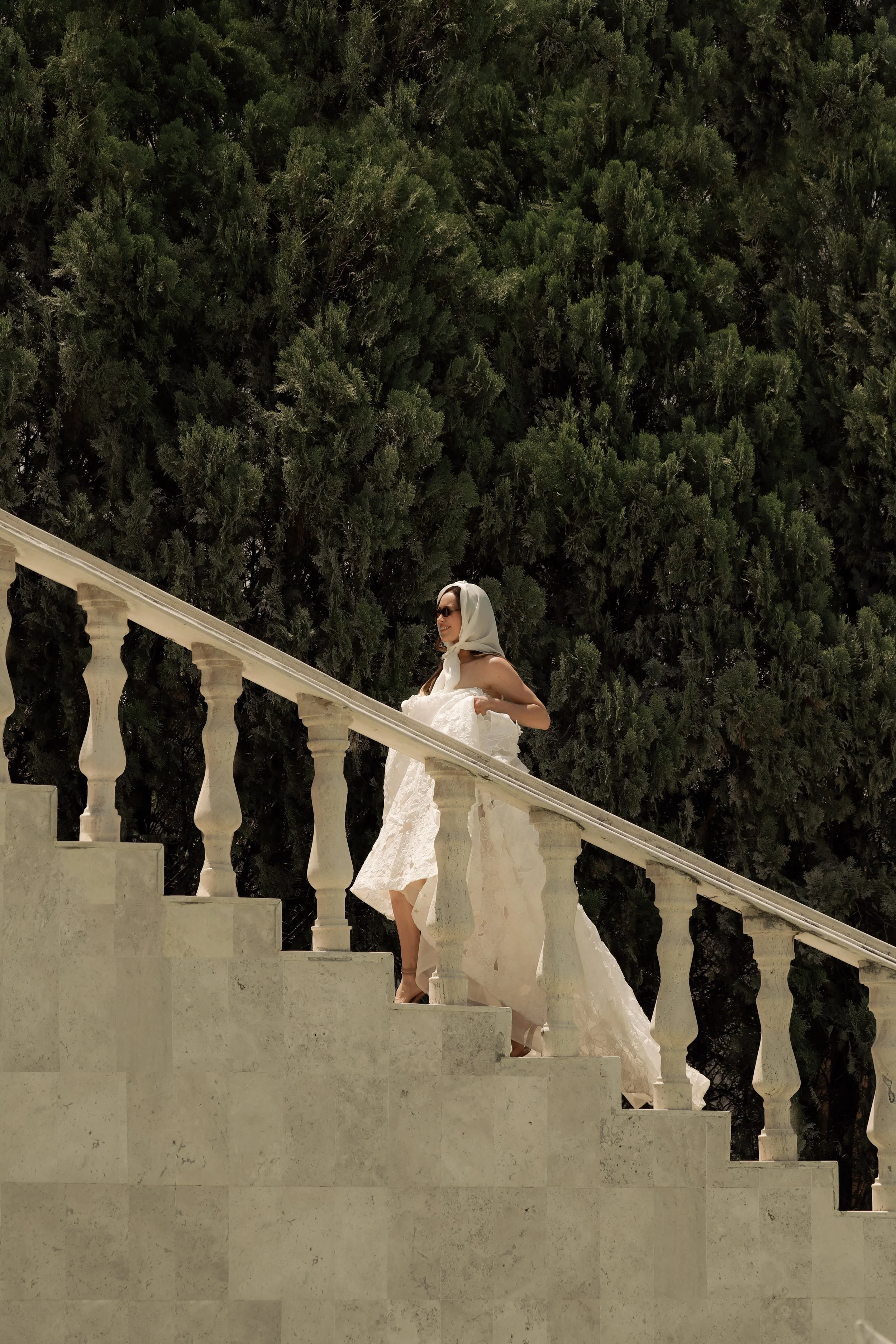 Guardiola  at New York Bridal Market. A woman in a wedding gown and sunglasses standing on a cream-colored stone staircase with a white balustrade, surrounded by tall green trees.