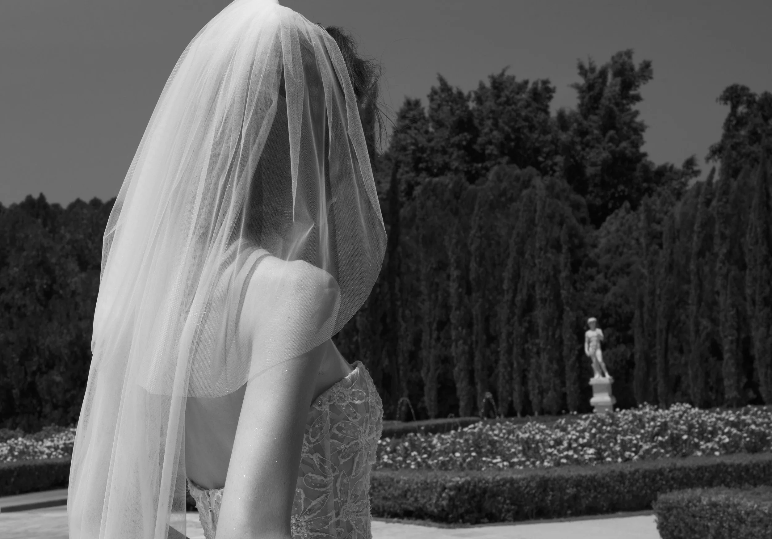 	
Guardiola at New York Bridal Market A woman in a lace dress with a veil over her head, outdoors in a park or garden, with trees and a statue in the background.