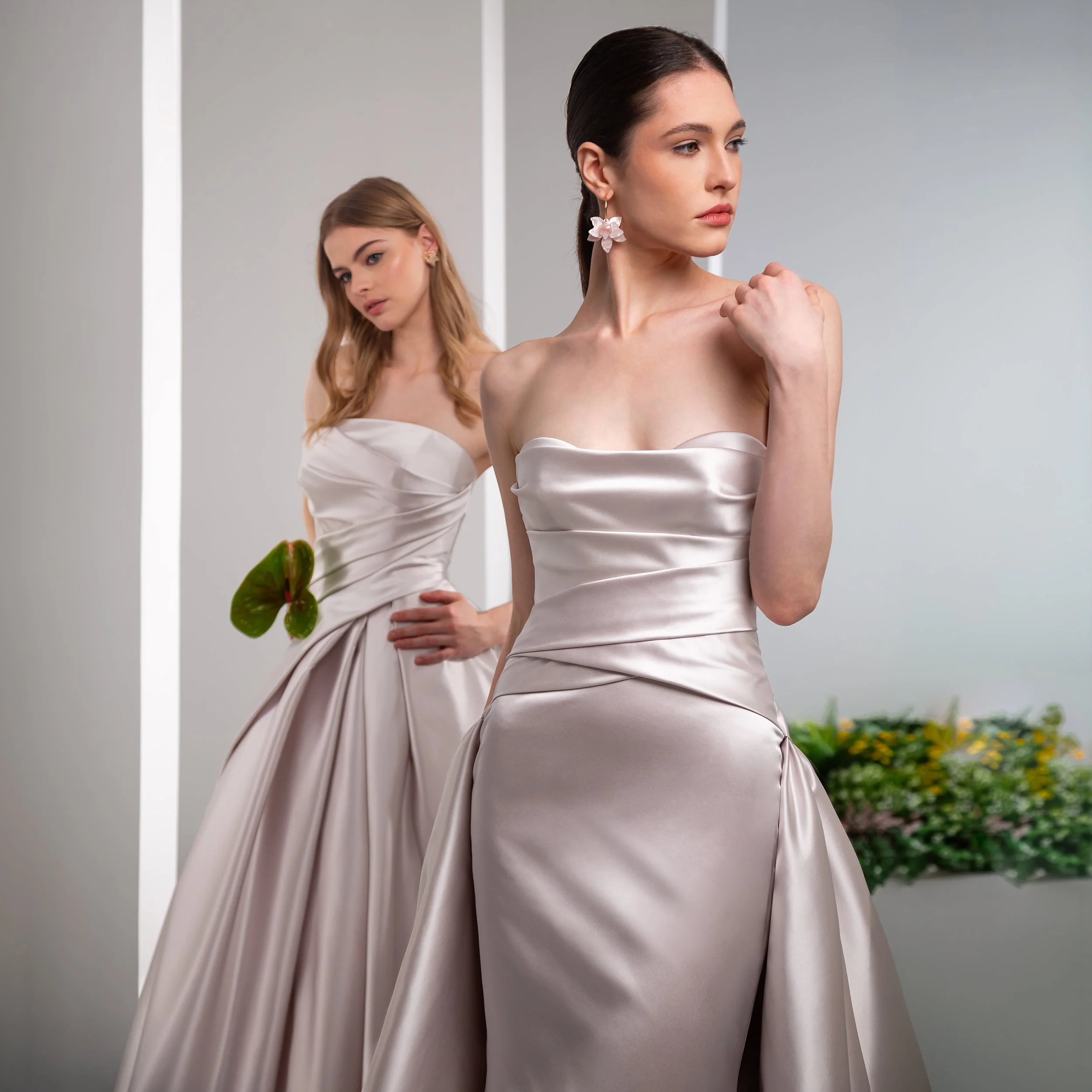 Valentini Spose at New York Bridal Market.    Two women dressed in satin gowns standing in front of a mirror, with one woman touching her shoulder and the other looking at her.