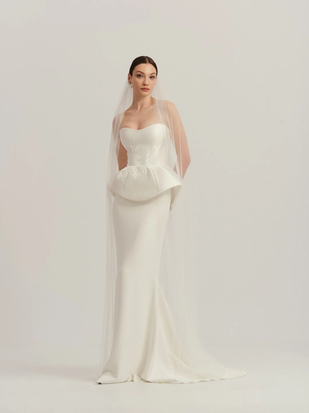 Divine Atelier at New York Bridal Market  A woman in a strapless white wedding gown and veil standing against a plain light-colored background.