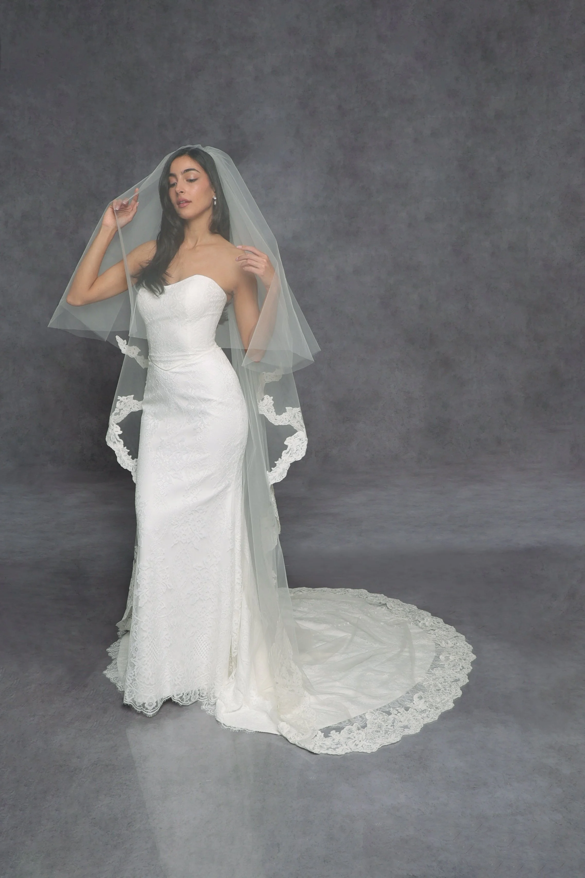 Robert Bullock at New York Bridal Market. A woman in a white wedding dress with lace details and an off-the-shoulder neckline, holding a sheer veil with lace trim, standing against a gray backdrop.