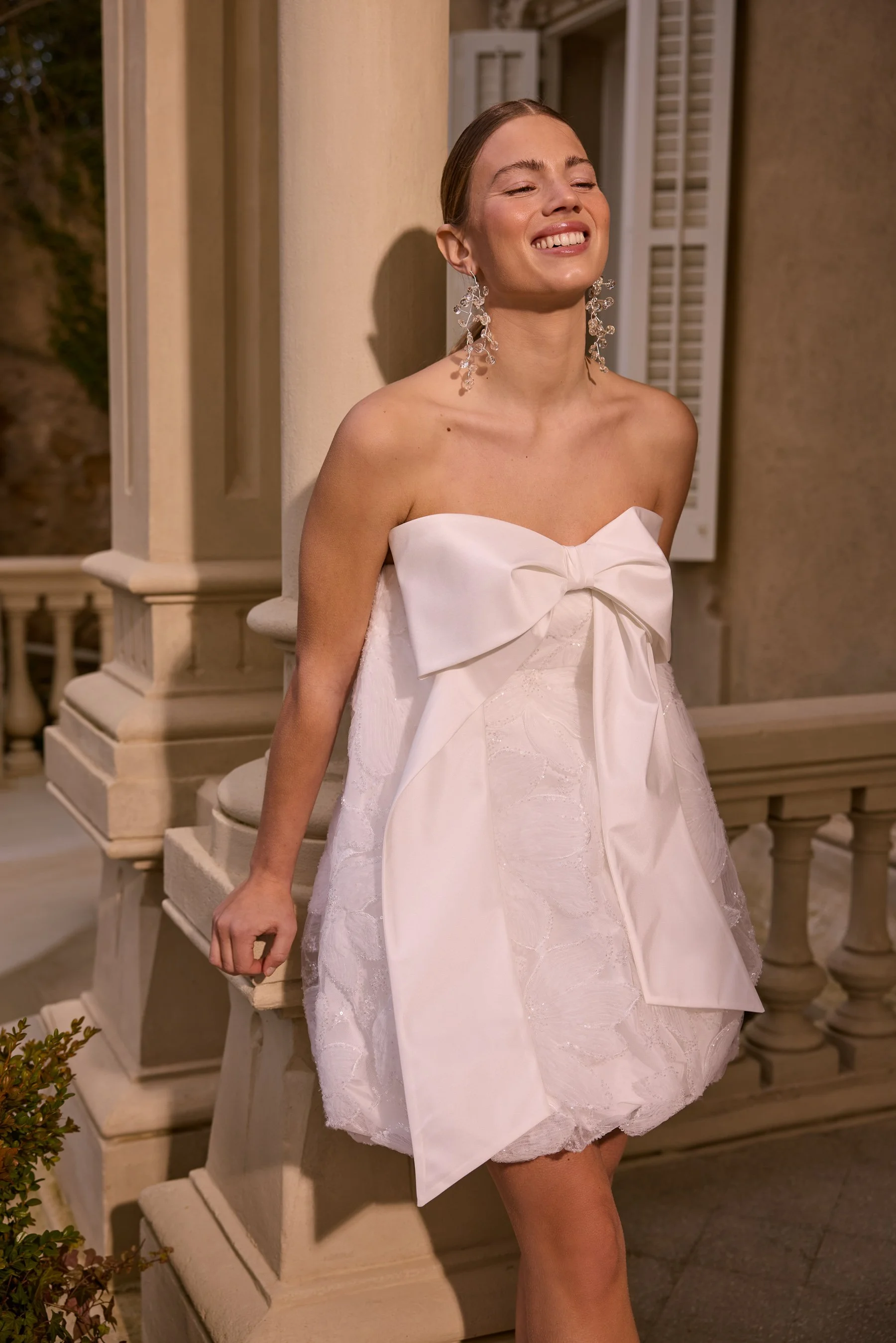 Jesus Peiro at New York Bridal Market.  A smiling woman in a white strapless dress with a large bow on the front, wearing large, ornate earrings, standing on a porch next to a balustrade.
