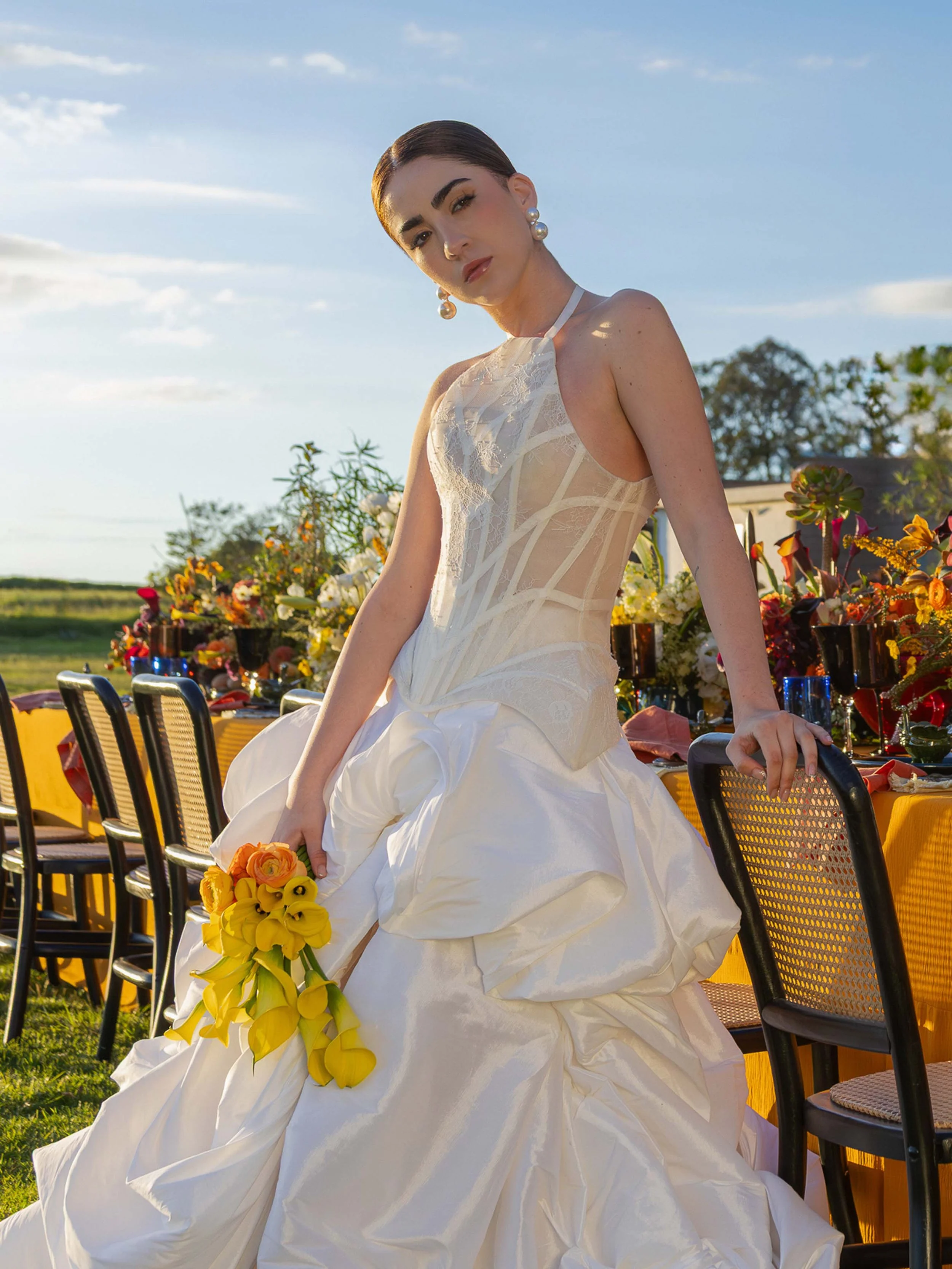 A woman in a white wedding gown sitting outdoors at sunset, holding a bouquet of yellow and orange flowers, with tables and chairs in the background decorated with flowers.