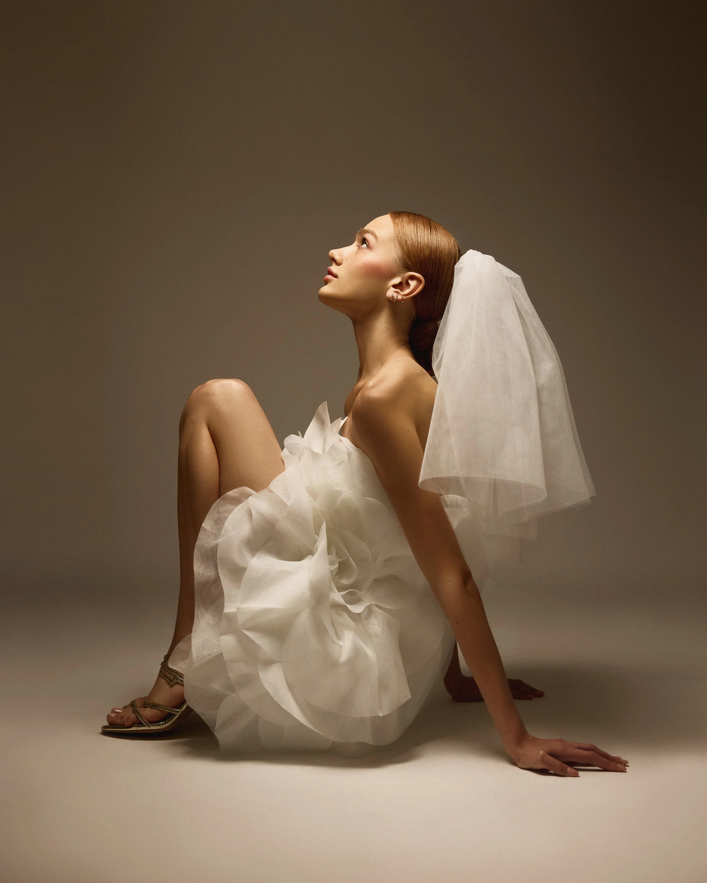 Gemy Maalouf at New York Bridal Makrket A woman sitting on the floor with one hand on the ground and her leg bent, wearing a white dress with ruffles and high heel sandals, against a plain background.