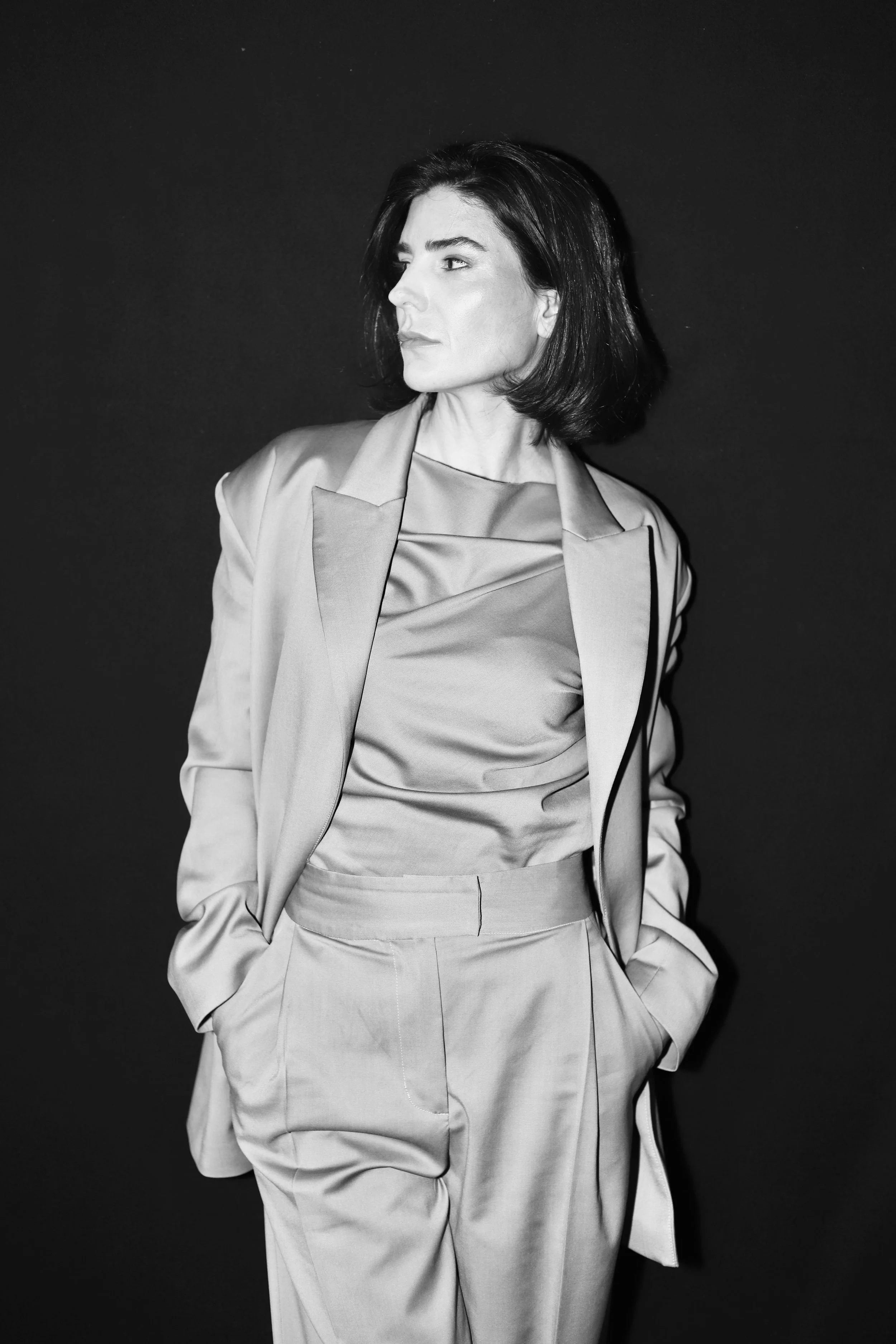 Sophie et Voila at New York Bridal Market. Black and white photo of a woman with shoulder-length dark hair, wearing a suit jacket and matching high-waisted pants, standing against a plain dark background, looking to her left.