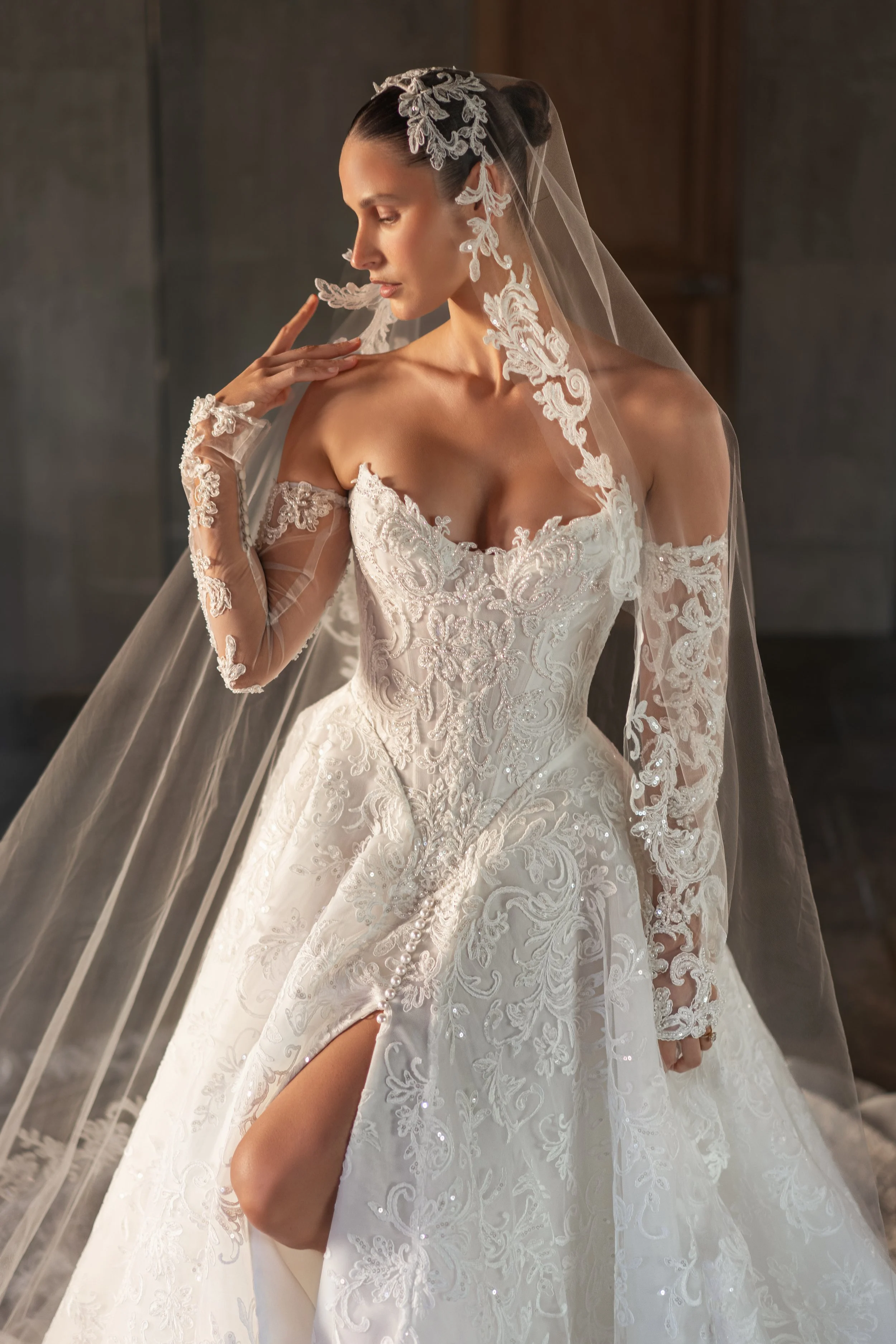 Blanche Bridal at New York Bridal Market  A bride wearing a white wedding gown with lace details and a long lace veil, touching her face with one hand and looking down.