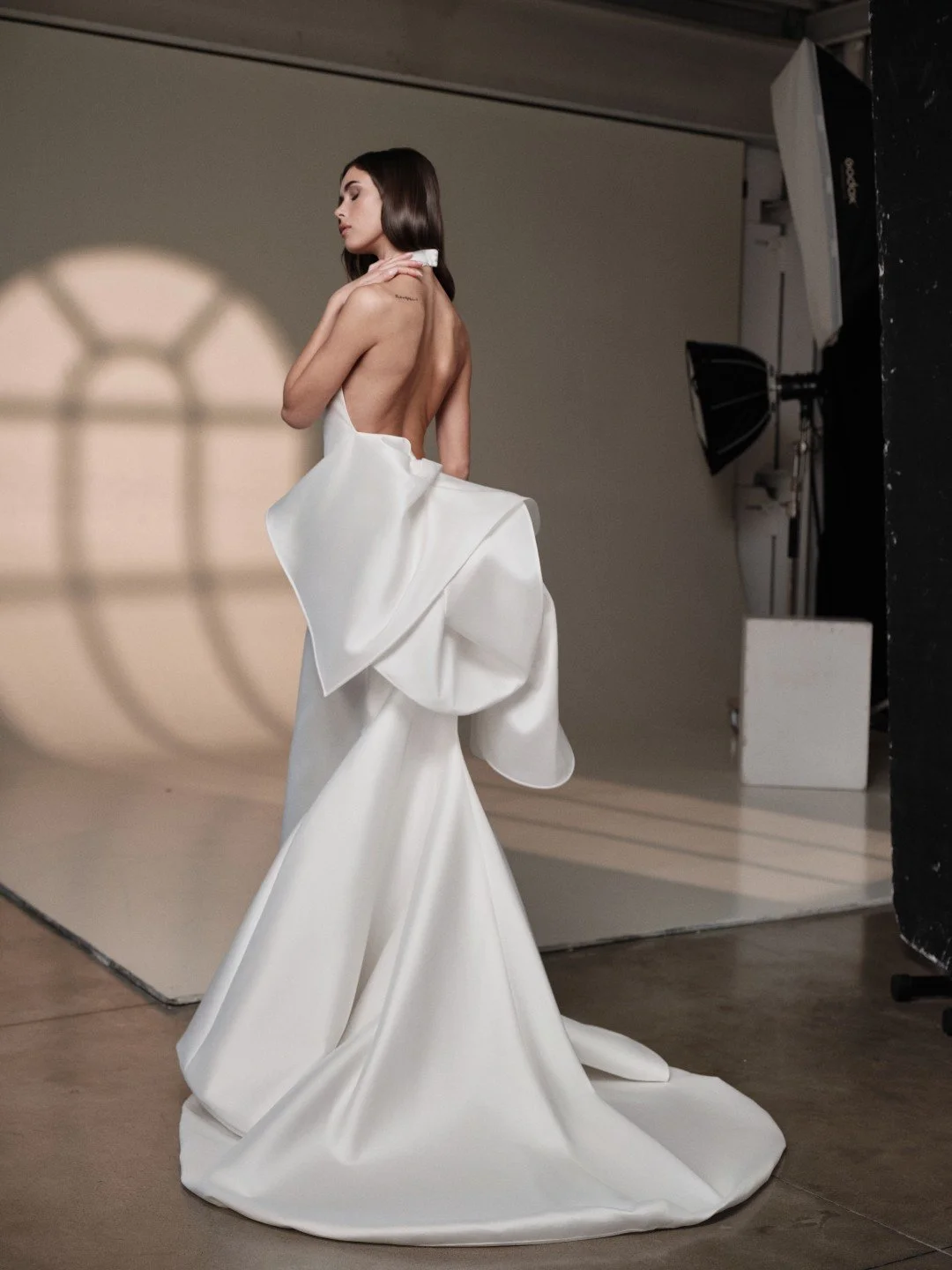 Maria Pia at New York Bridal Market.  A woman in a white strapless gown with a large bow detail at the back posing in a photography studio with studio equipment and a shadow of a window on the wall.