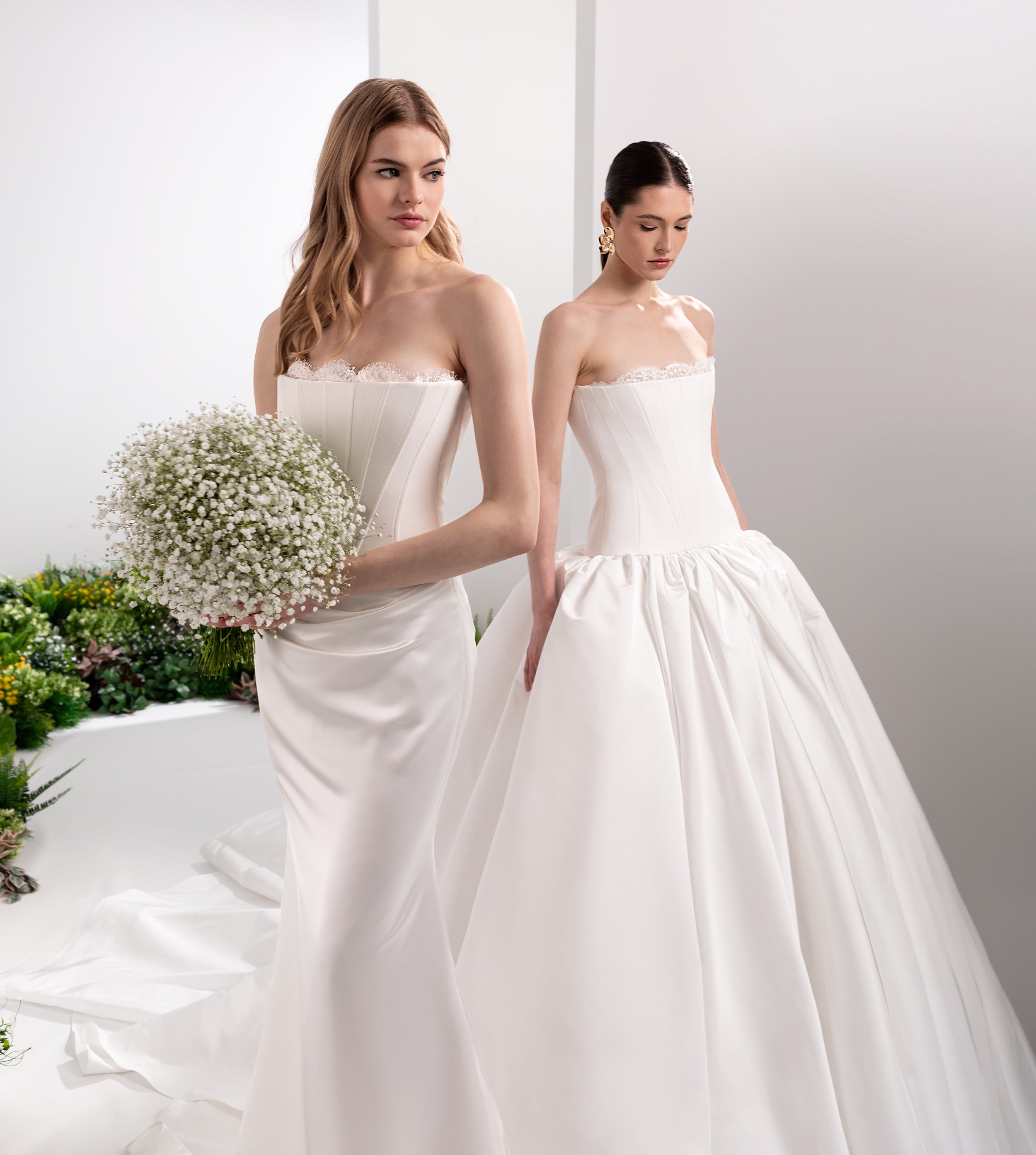 Valentini Spose at New York Bridal Market.  Two women in white wedding dresses standing indoors, with a white background and greenery on the side. One woman is holding a bouquet of small white flowers.
